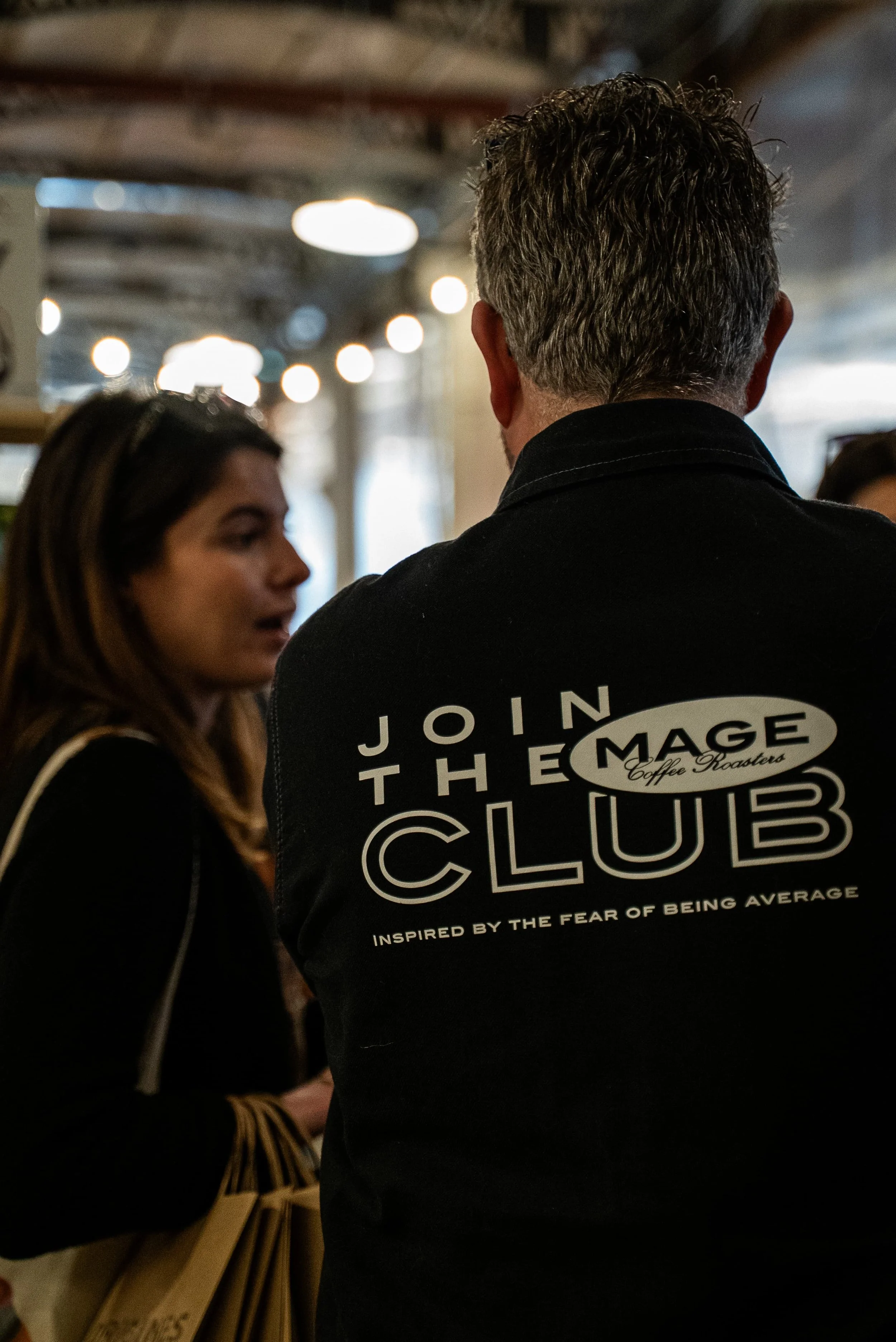 Un homme avec des cheveux gris portant un t-shirt noir avec un logo "Join the Amazing Club" vu de derrière, en conversation avec une jeune femme dans un lieu intérieur.