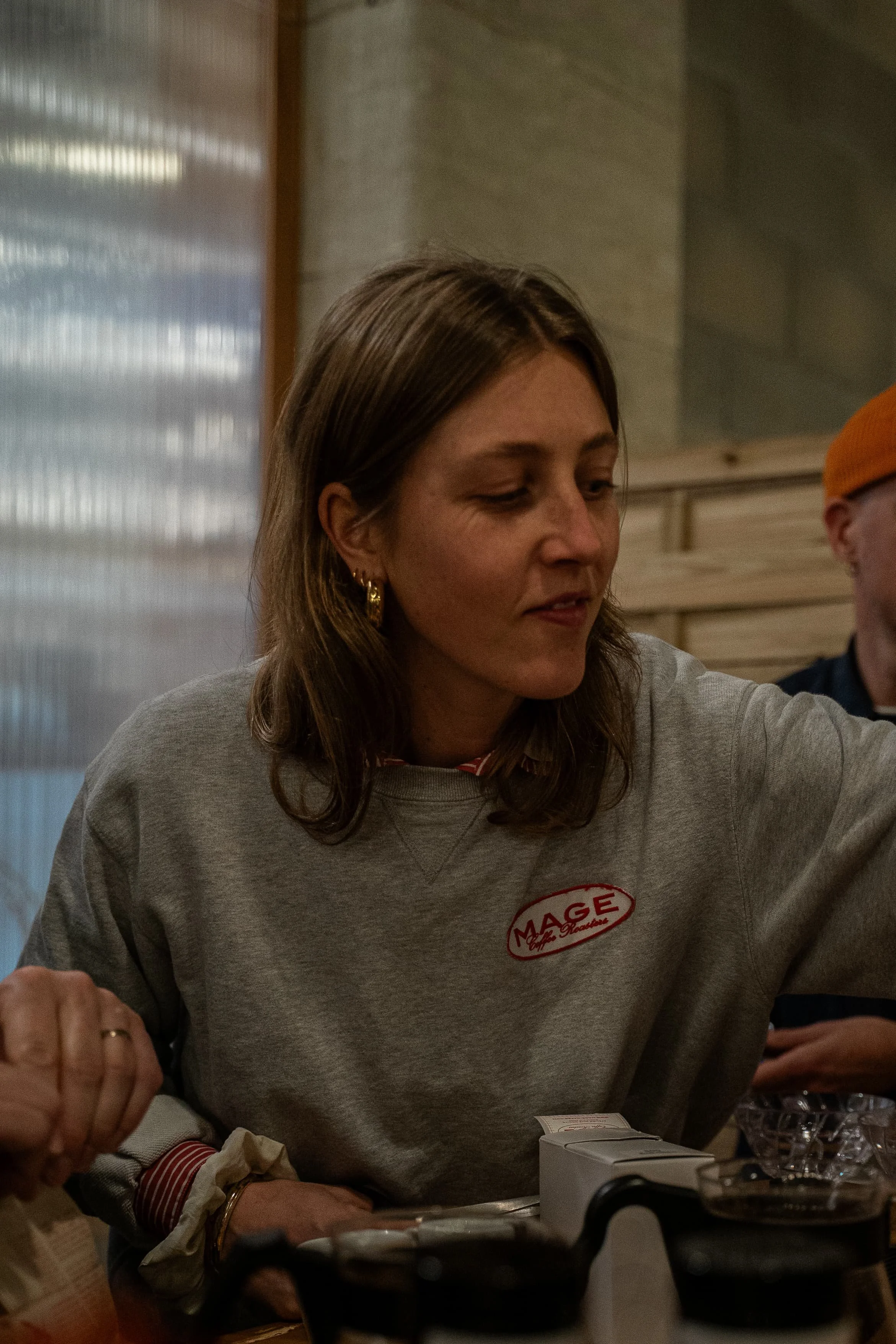 Une femme portant un sweatshirt gris avec un logo « MAGE » est assise à une table, entourée d'objets divers, peut-être en train de manger ou de discuter.