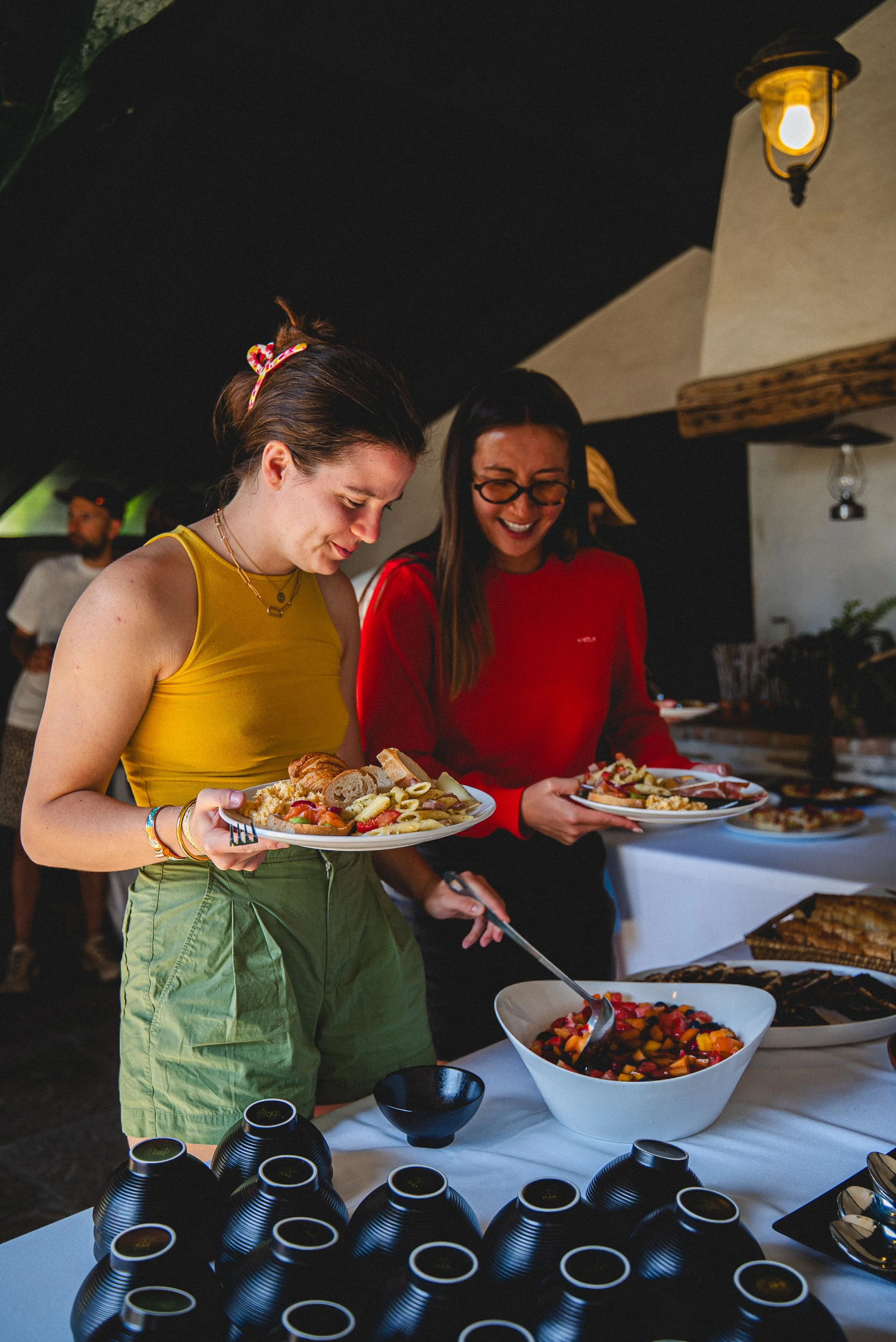 Deux femmes souriantes se servent de la nourriture lors d'un buffet, avec des bols de salade de fruits en premier plan.
