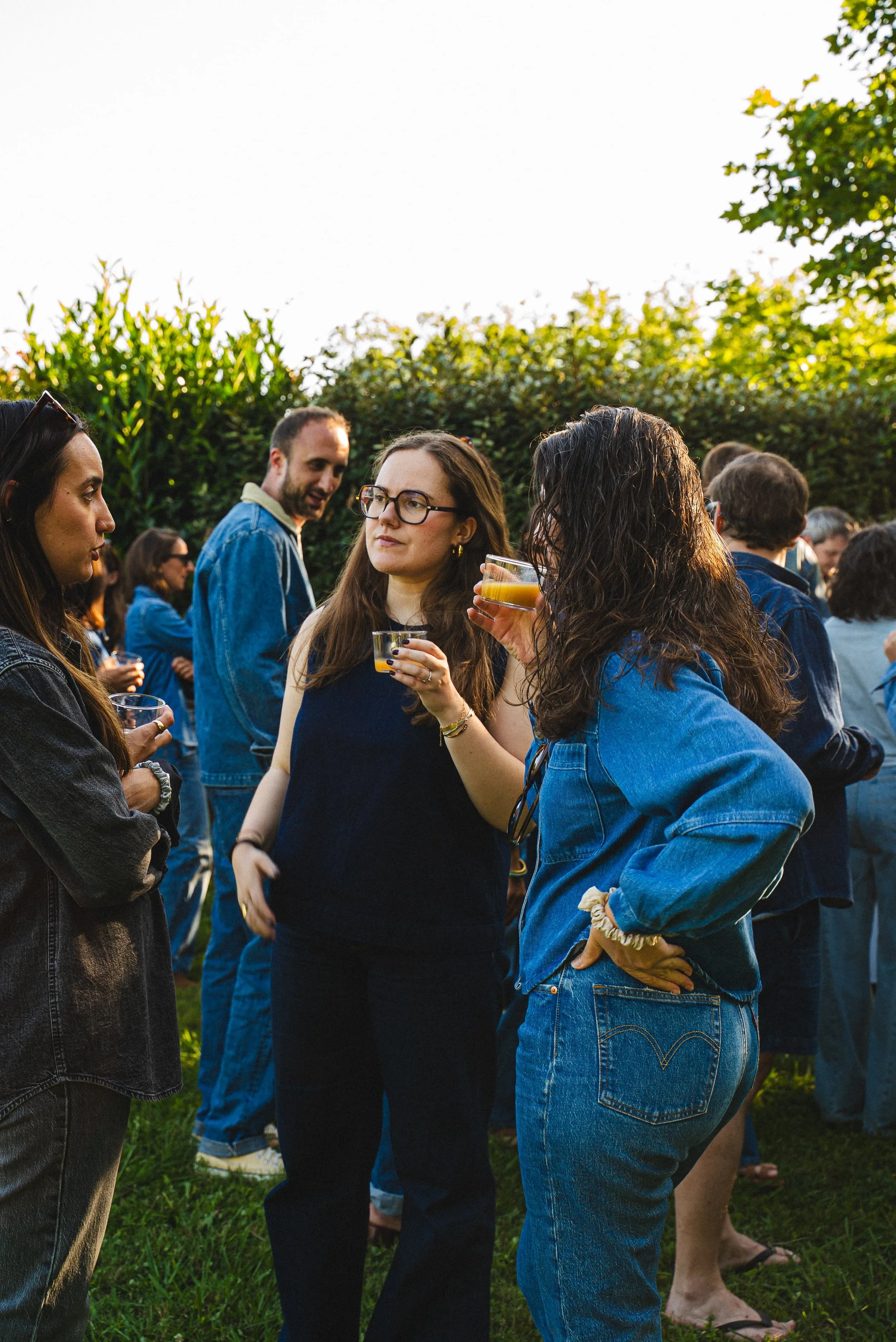 Groupe de personnes en conversation lors d'une réunion en plein air, en cercle dans un jardin avec des buissons en arrière-plan, certains tenant des boissons.