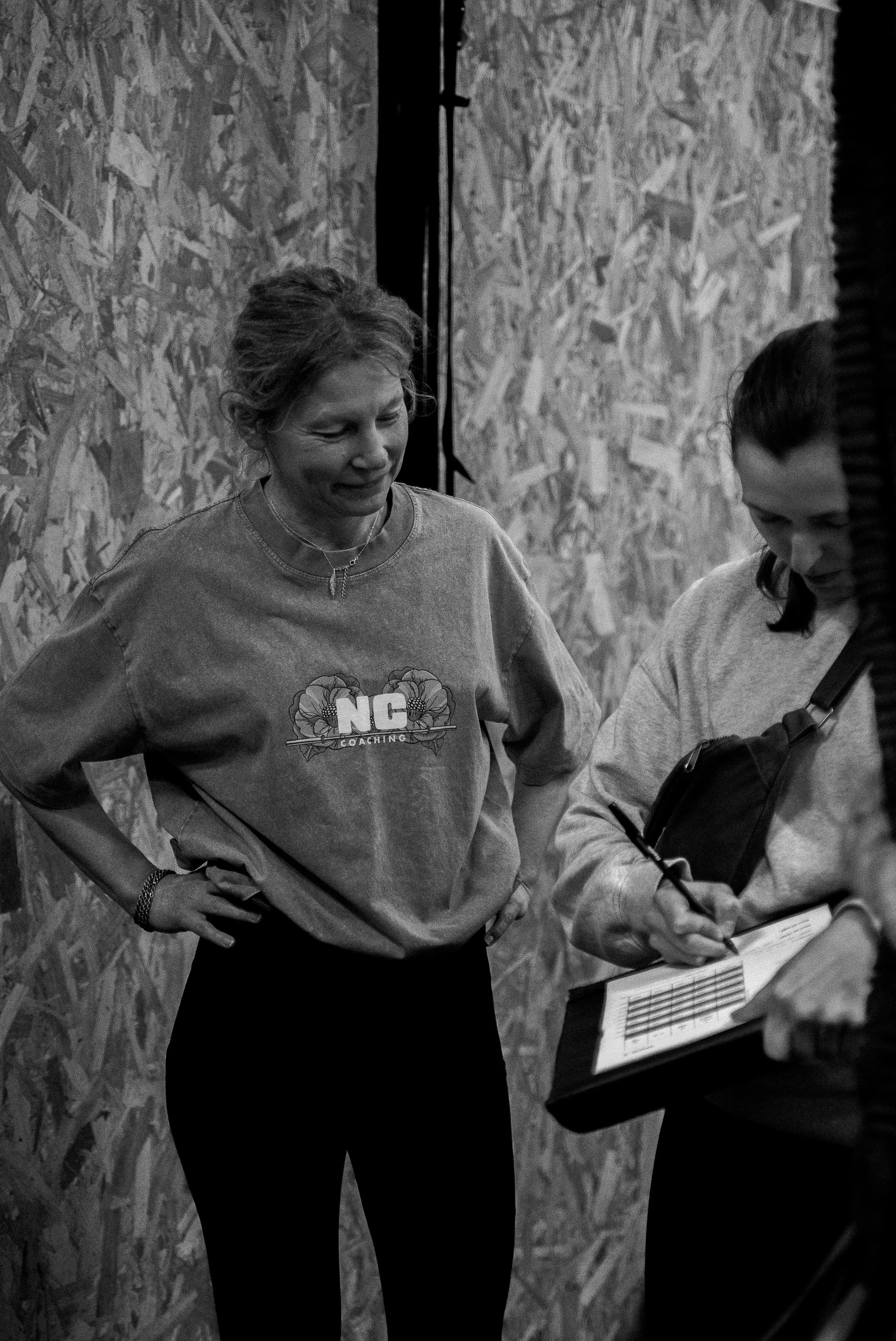 Deux femmes discutent devant un panneau d'information, l'une porte un sweat à logo NC Coaching et l'autre tient un carnet.