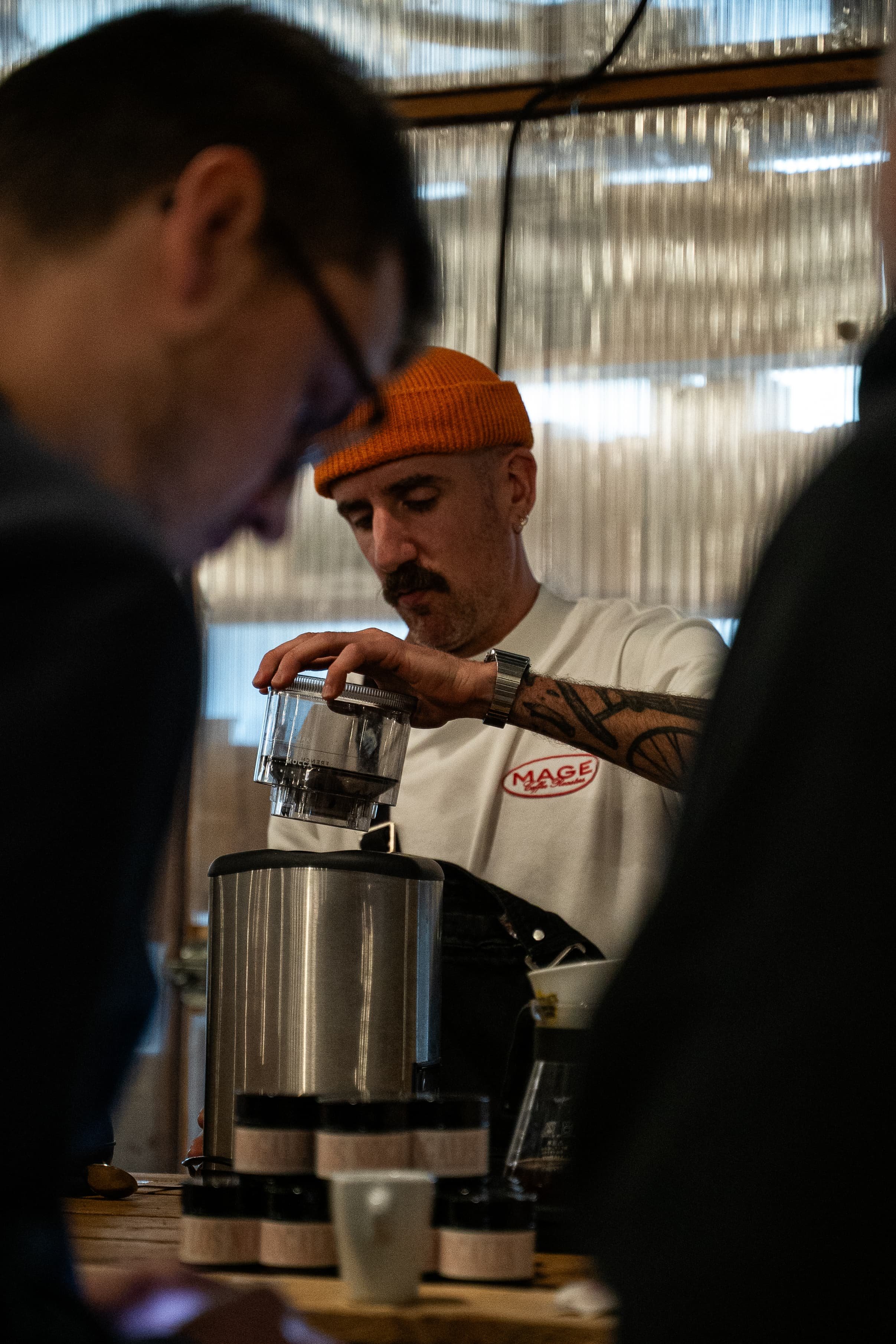 Un homme portant un bonnet orange et une chemise blanche avec un logo, utilisant une machine pour préparer une boisson, entouré d'autres personnes dans un environnement intérieur.