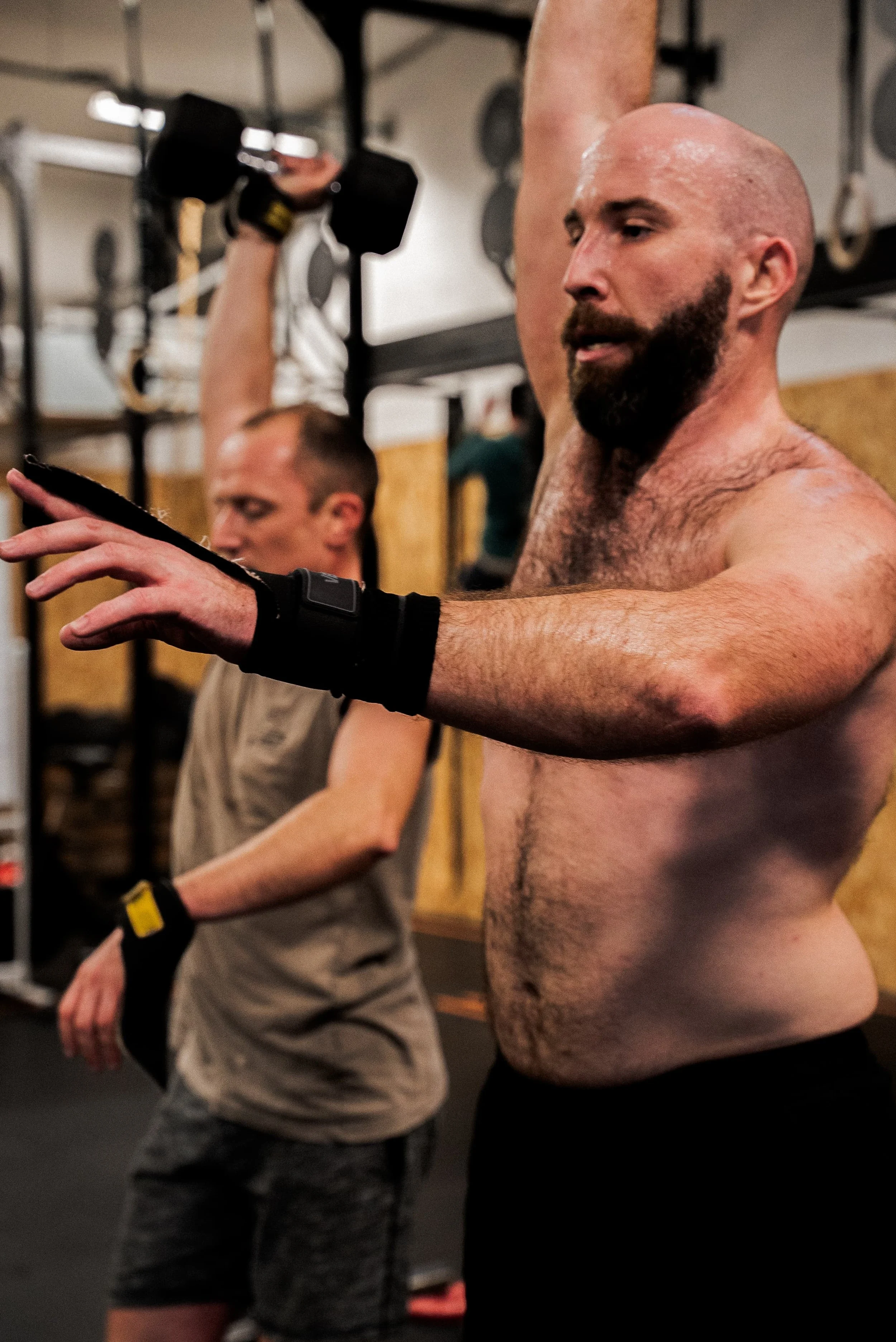 Deux hommes s'entraînent dans une salle de sport. L'un d'eux, sans shirt, fait un exercice au bras avec des haltères, tandis que l'autre, porte un t-shirt gris et des gants de musculation, se trouve à côté.