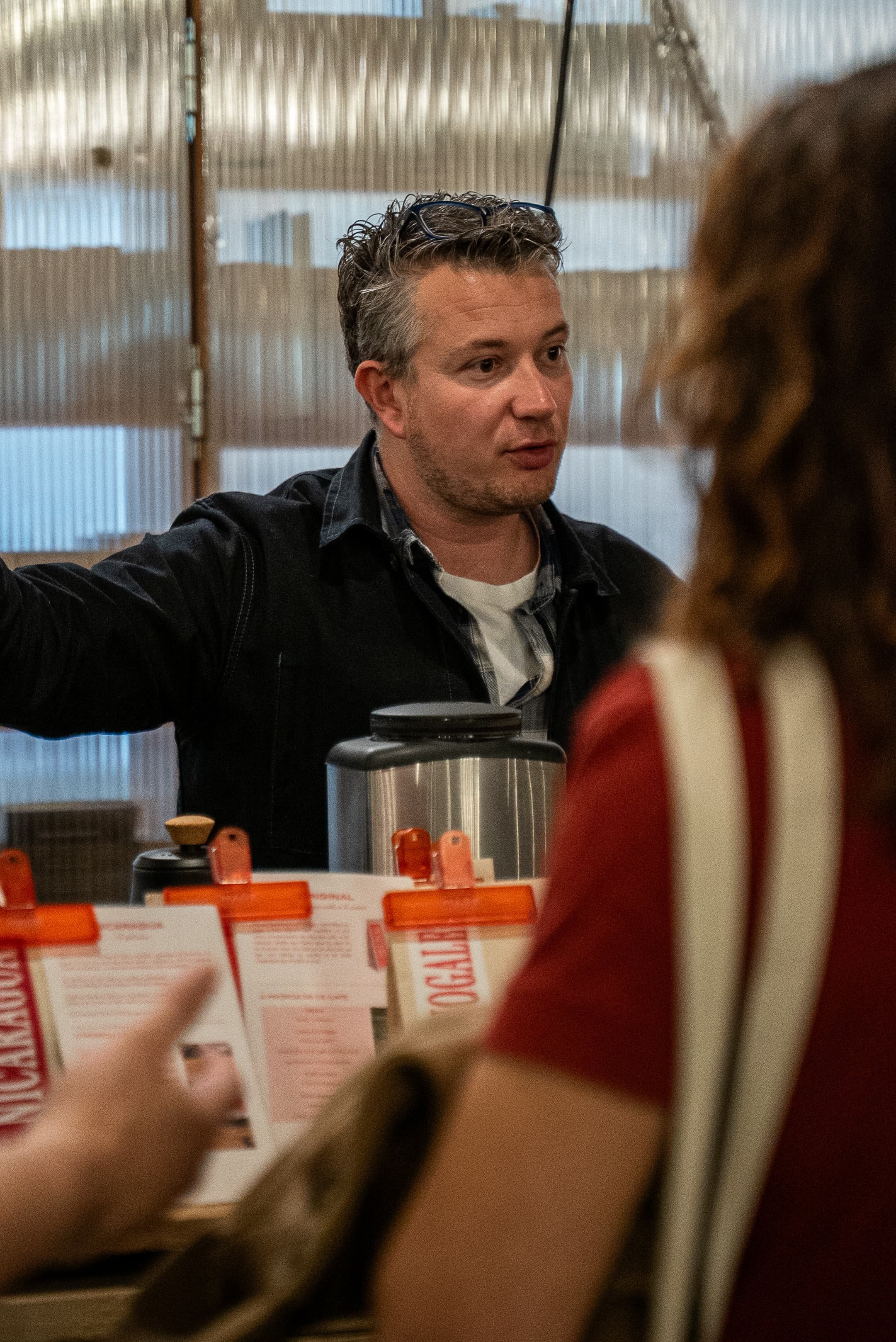 Un homme discute avec une fille dans un lieu intérieur, peut-être un café ou un restaurant, avec des rayonnages en bois, et des menus ou des flyers à l'avant-plan.