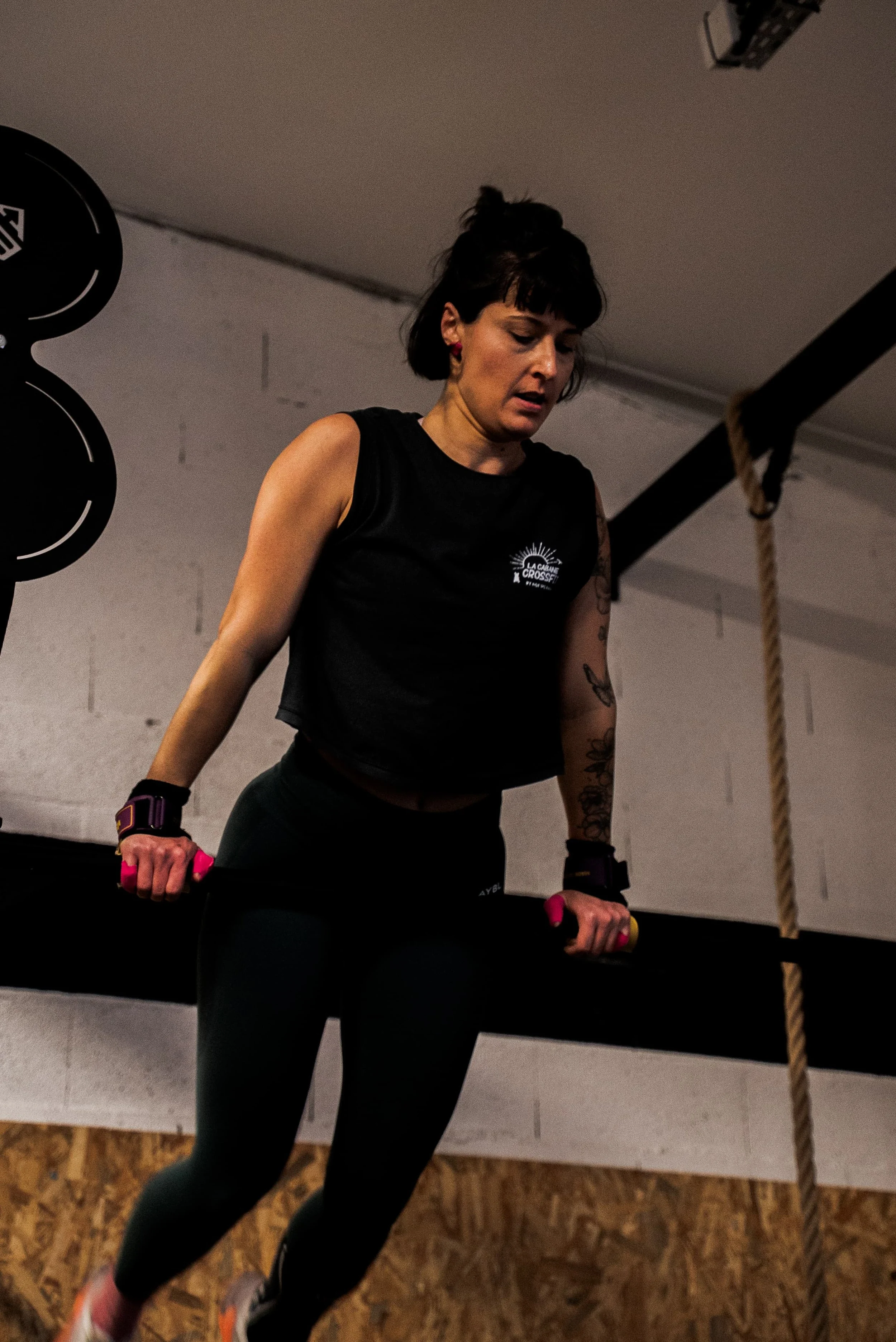 Une femme en vêtements de sport, tenant des haltères roses et noirs, faisant de l'exercice dans une salle de sport. Elle porte un débardeur noir avec un logo "La Cabane Crossfit".