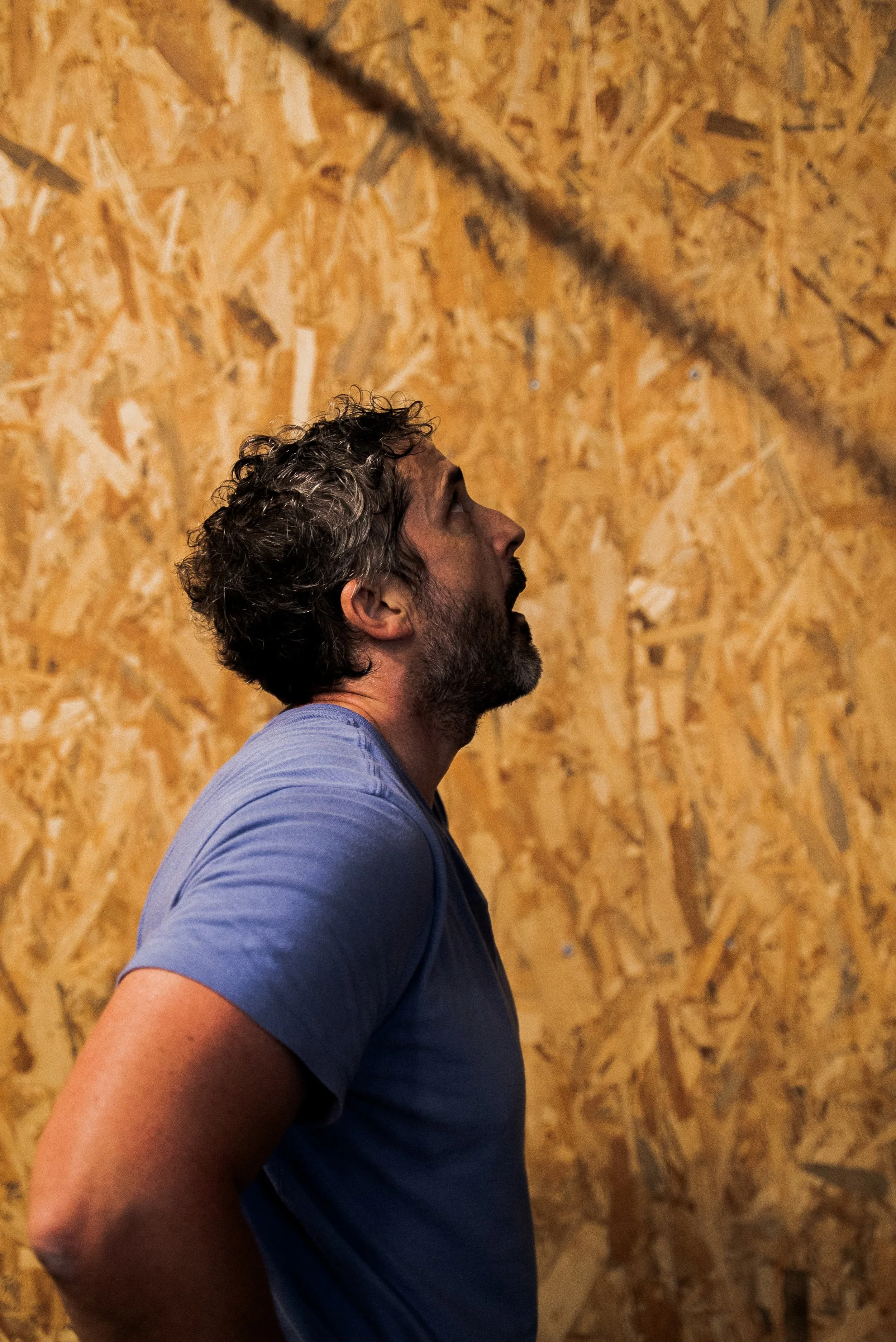 Un homme à barbe et cheveux bouclés regarde vers le haut, portant un t-shirt bleu, avec un mur en bois derrière lui.