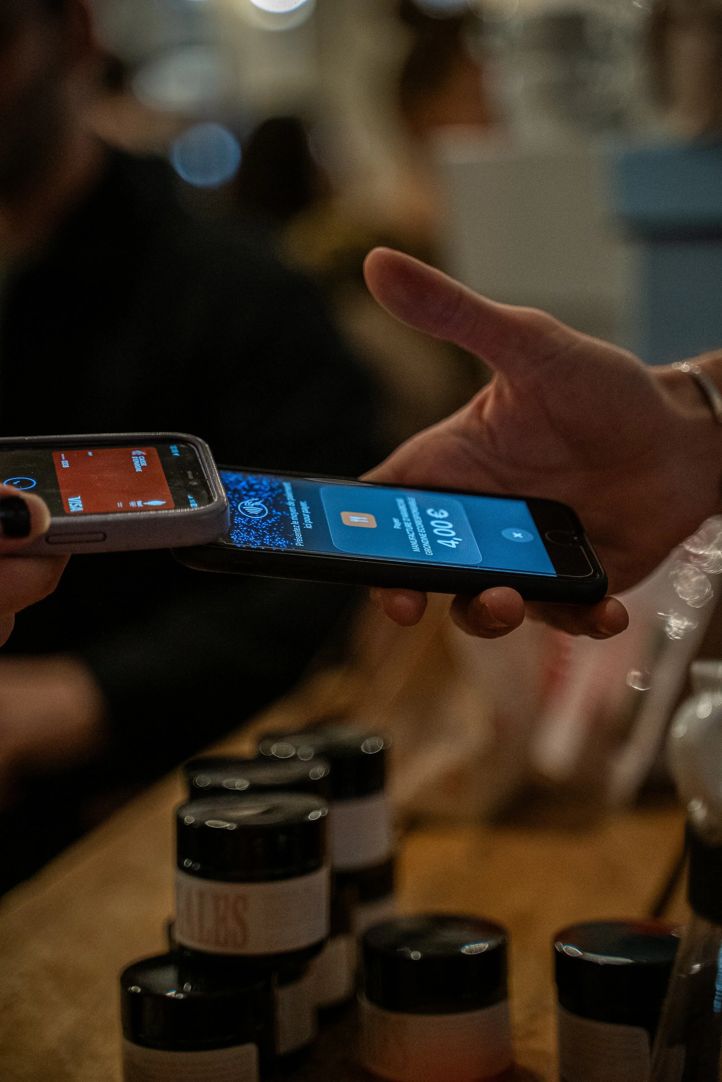 Une main tient un téléphone portable affichant un prix de 4,06 €, avec un fond flou de bouteilles de médicaments ou de compléments alimentaires.