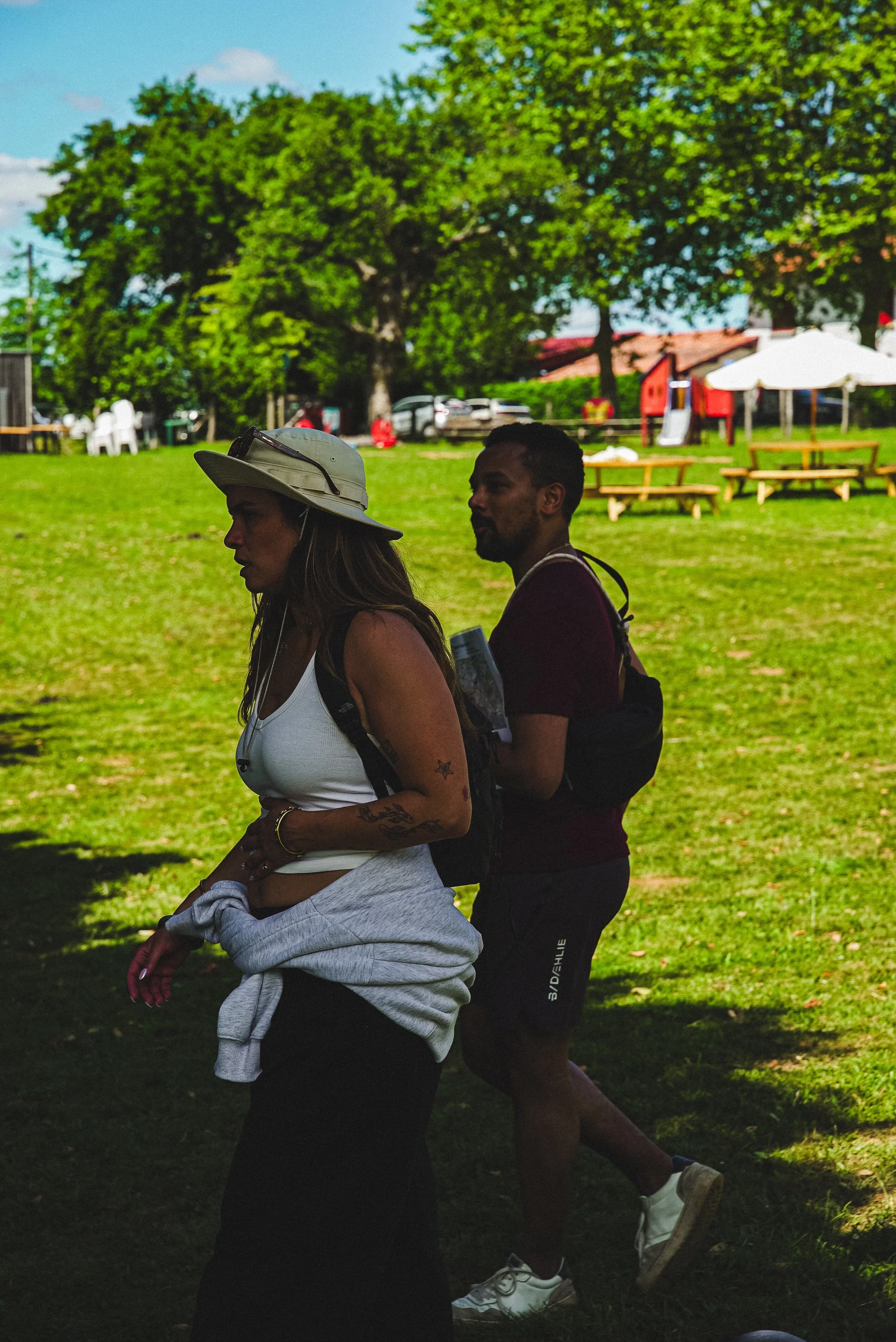 Deux personnes marchant dans un parc en plein jour, avec des arbres verts en arrière-plan et des bancs en bois sur l'herbe.