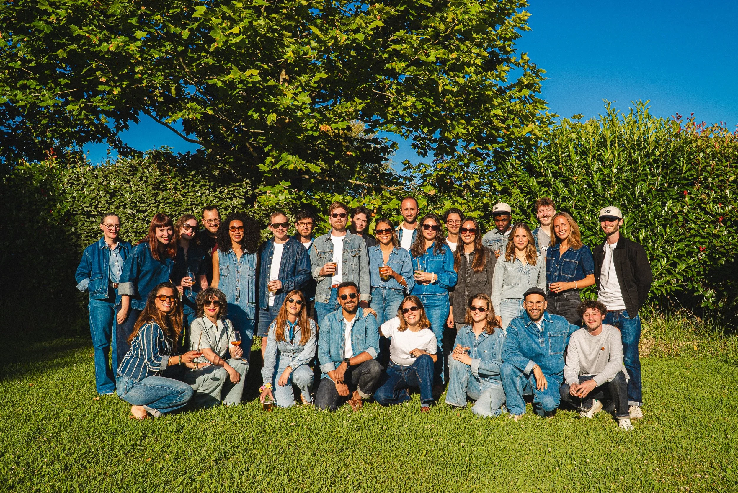 Groupe de personnes posant pour une photo en extérieur, certains tenant des verres, tous portant des vêtements décontractés, devant une haie et un arbre avec un ciel bleu.
