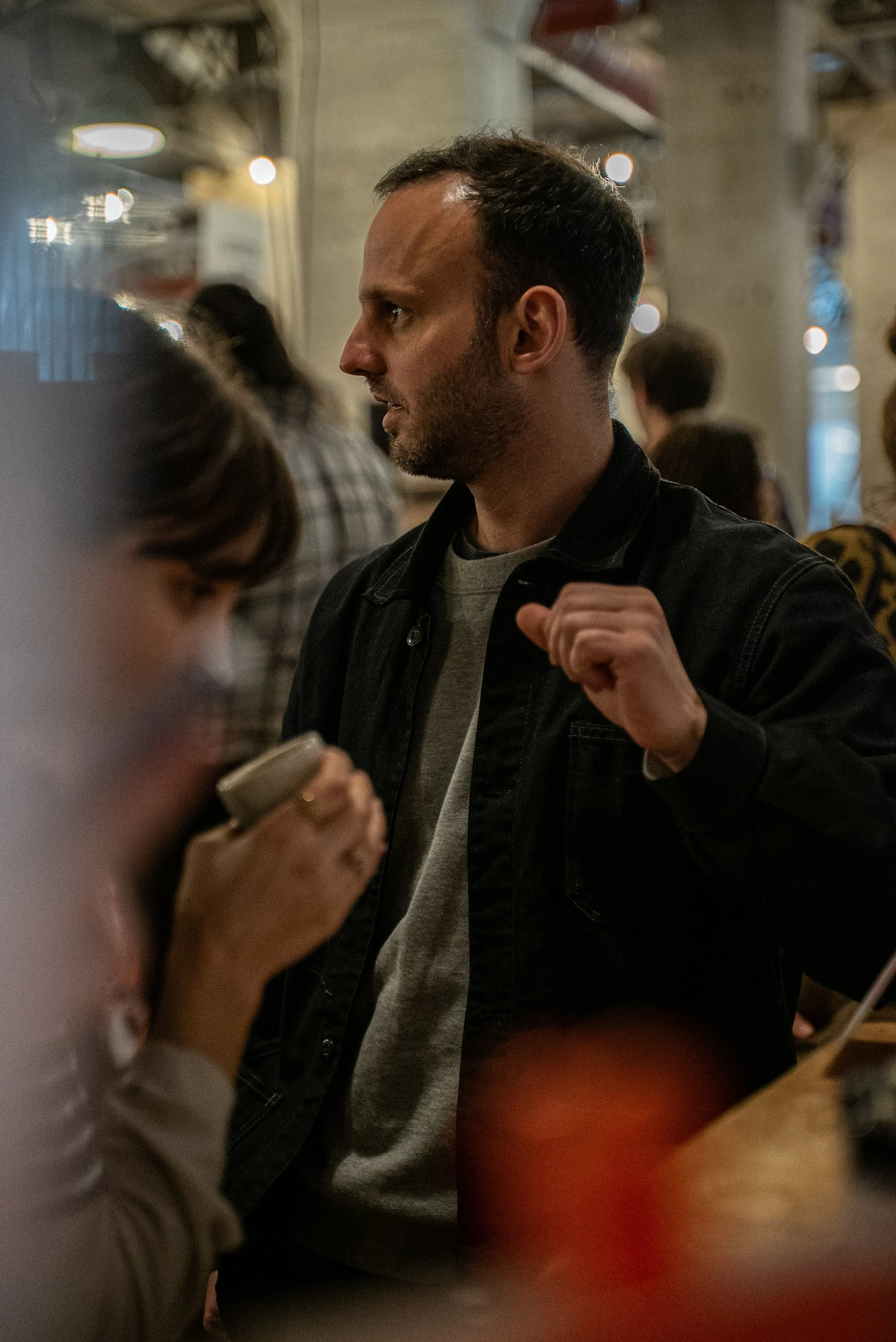 Un homme avec une barbe et des cheveux courts, portant un pull gris et une veste noire, est dans une ambiance intérieure avec d'autres personnes en arrière-plan. Il semble concentré ou en conversation, dans un espace probablement public comme un café