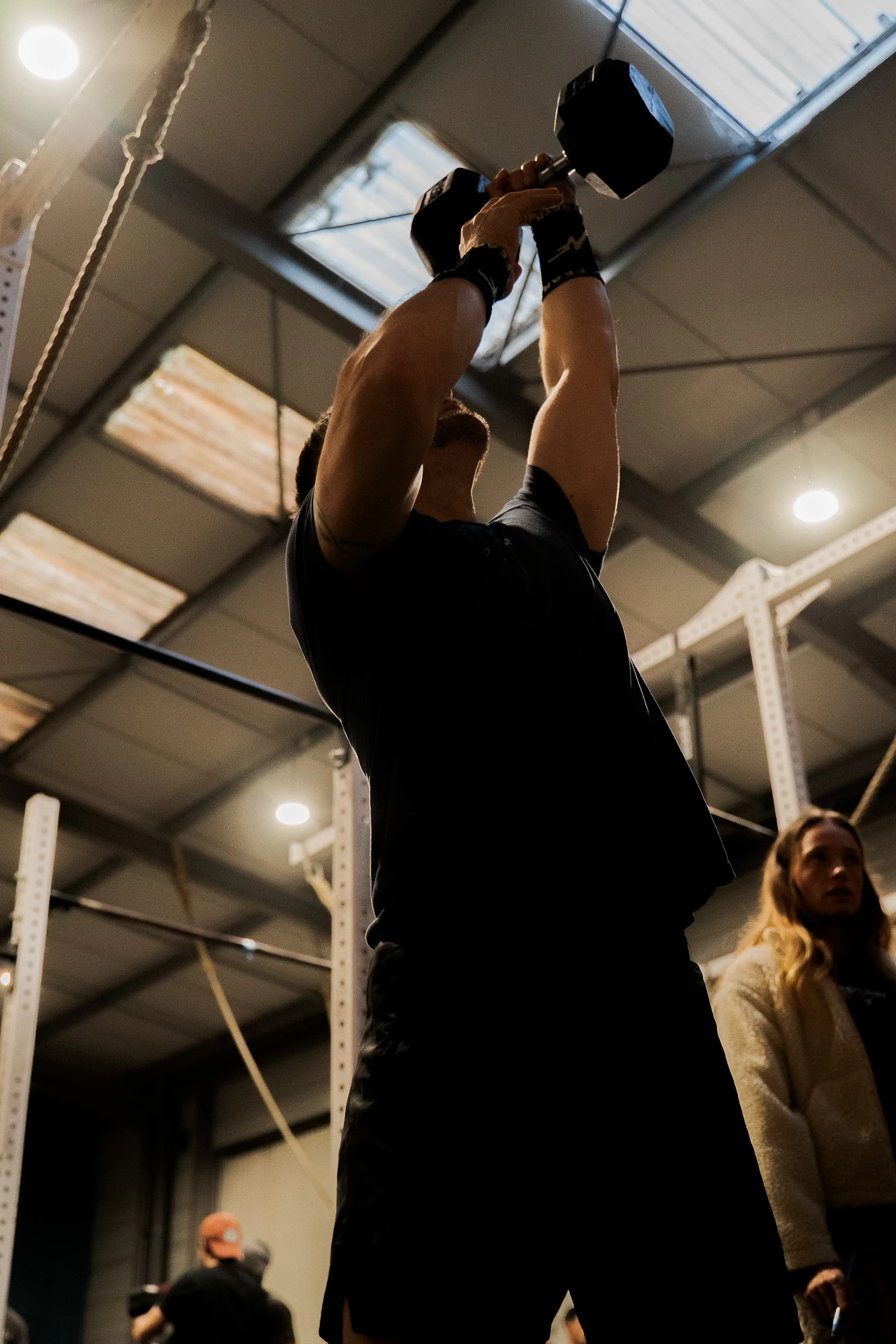 Une personne soulève un haltère dans une salle de sport, vue partiellement de dessous. Il y a d'autres personnes en arrière-plan, dont un homme avec des lunettes noires et une femme avec des cheveux blonds, dans un environnement intérieur avec des pl