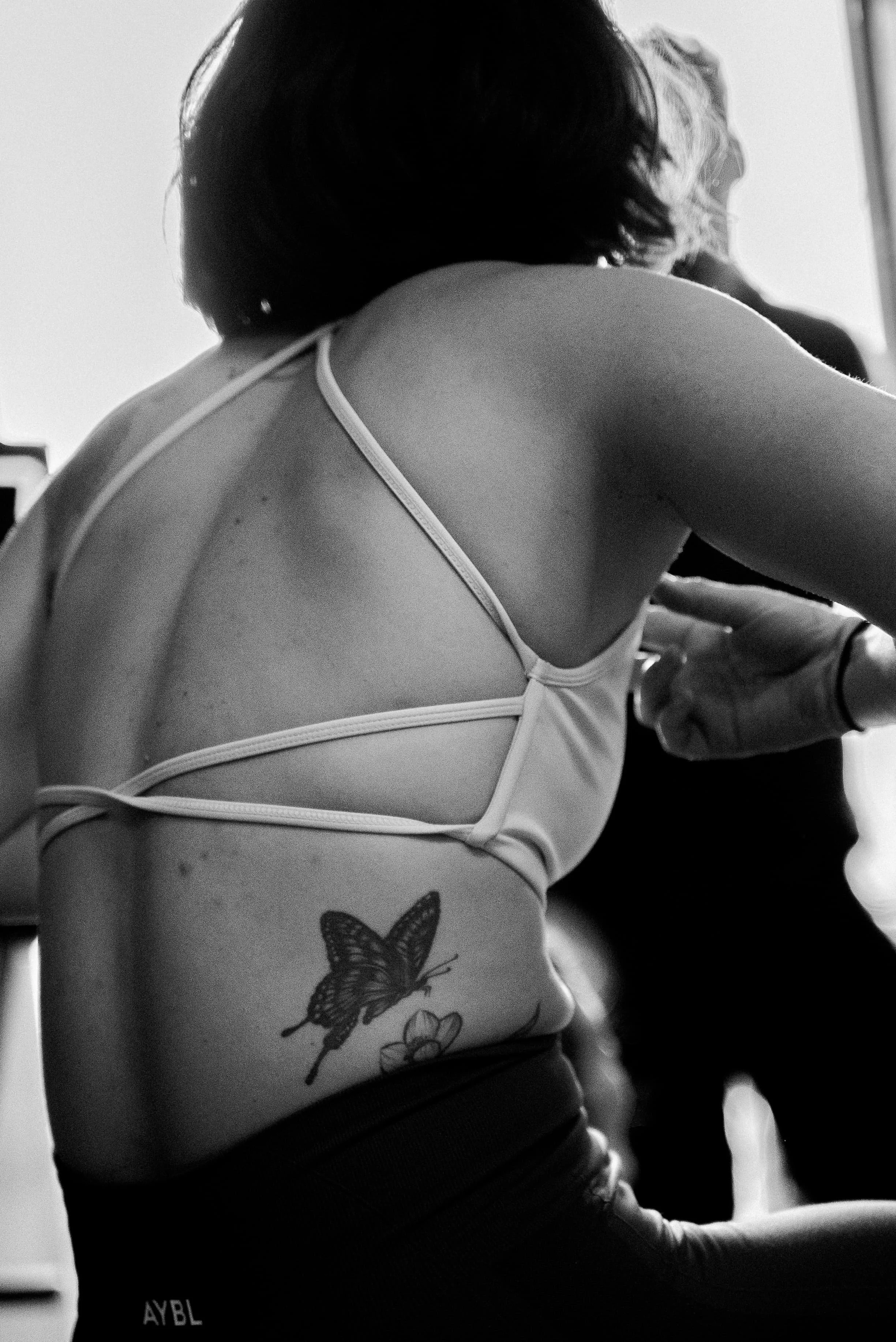 Vue de profil d'une femme de dos portant un débardeur avec des tatouages de papillons, dans une pièce lumineuse.