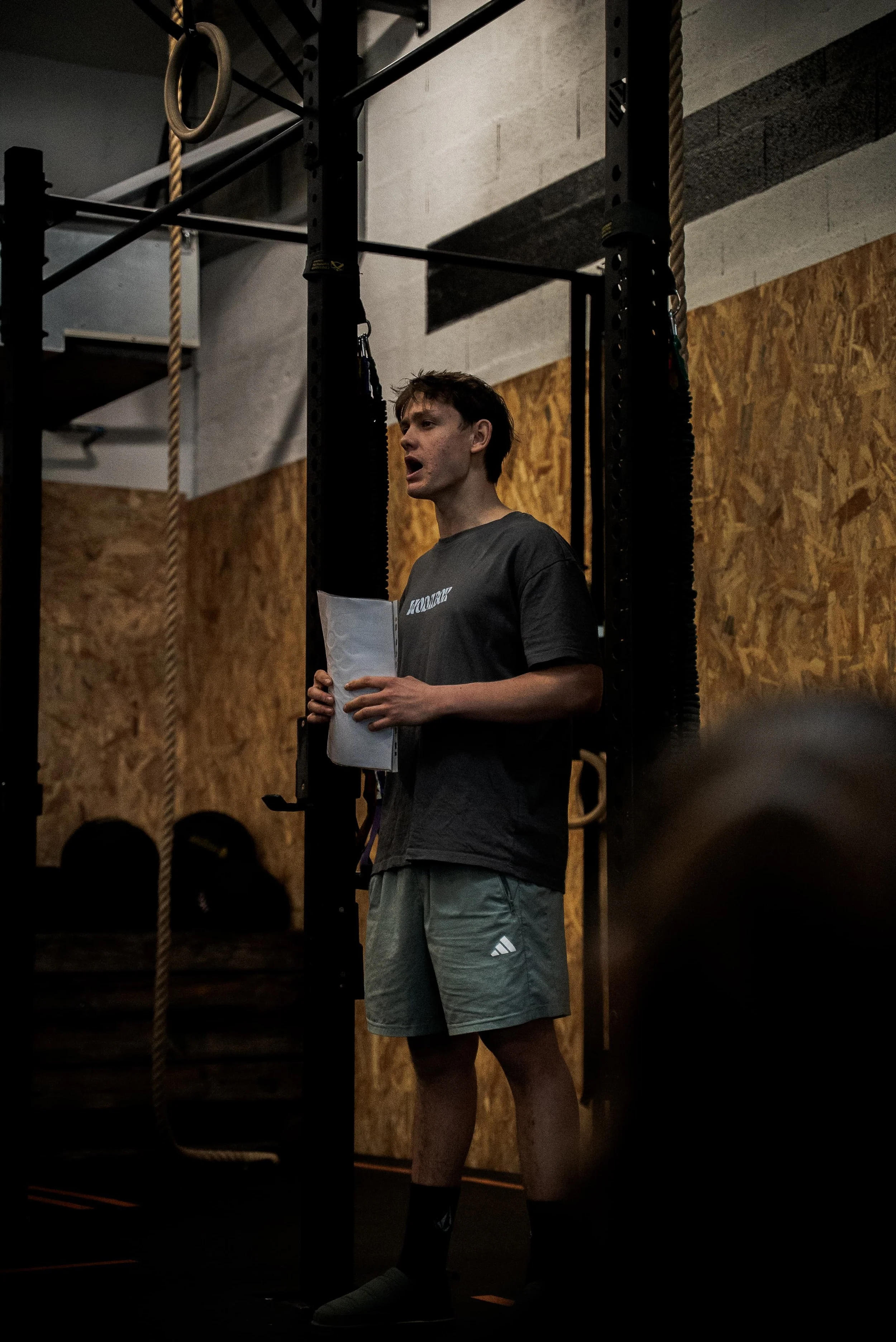 Jeune homme en salle de sport, vêtu d'un t-shirt gris et d'un short gris, tenant une feuille de papier, entouré de matériel d'entraînement, dans une ambiance sombre et concentrée.