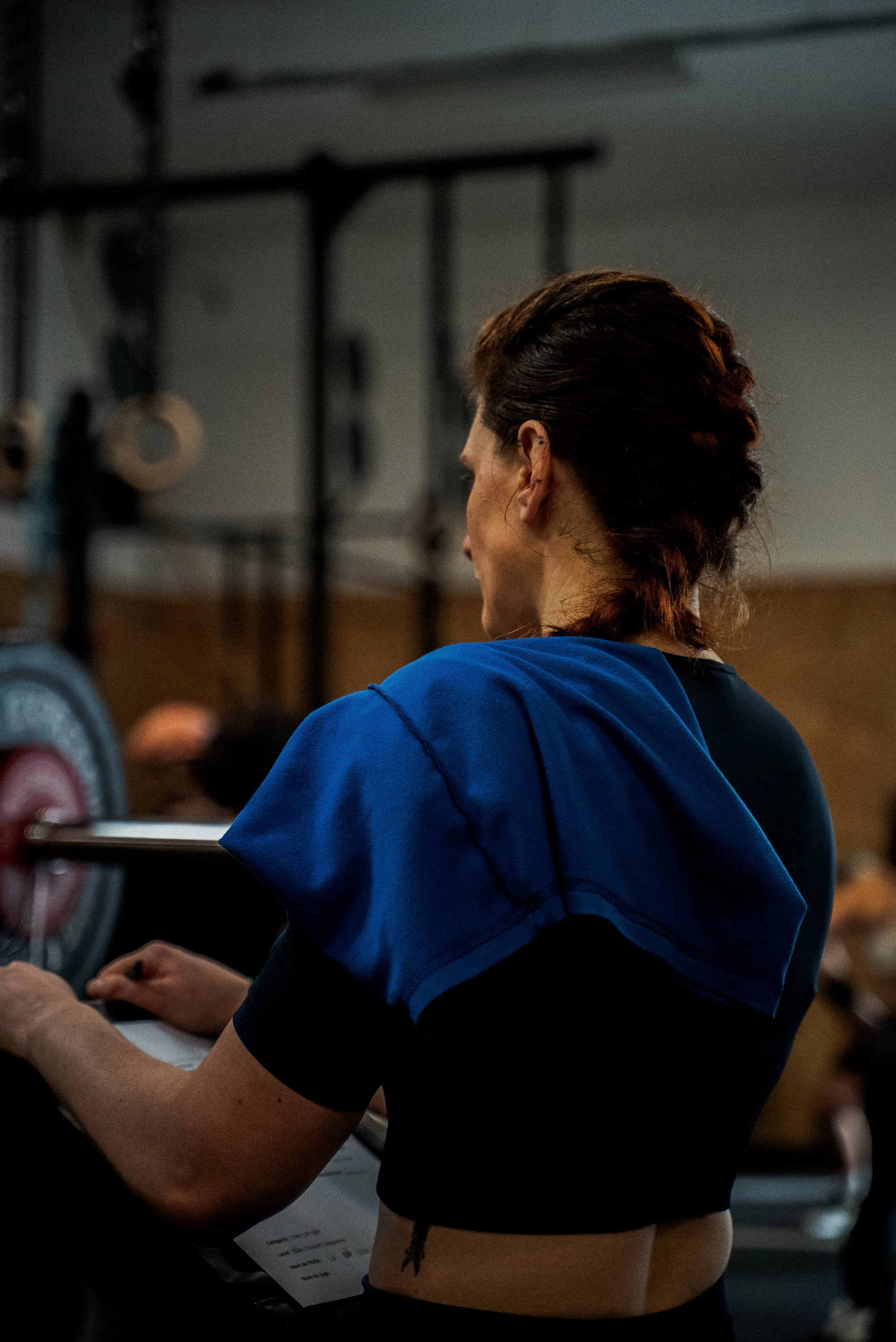 Une femme avec des cheveux bruns en natte, portant un haut de sport noir et un sweat bleu, regarde vers la droite lors d'une séance d'entraînement dans une salle de sport.