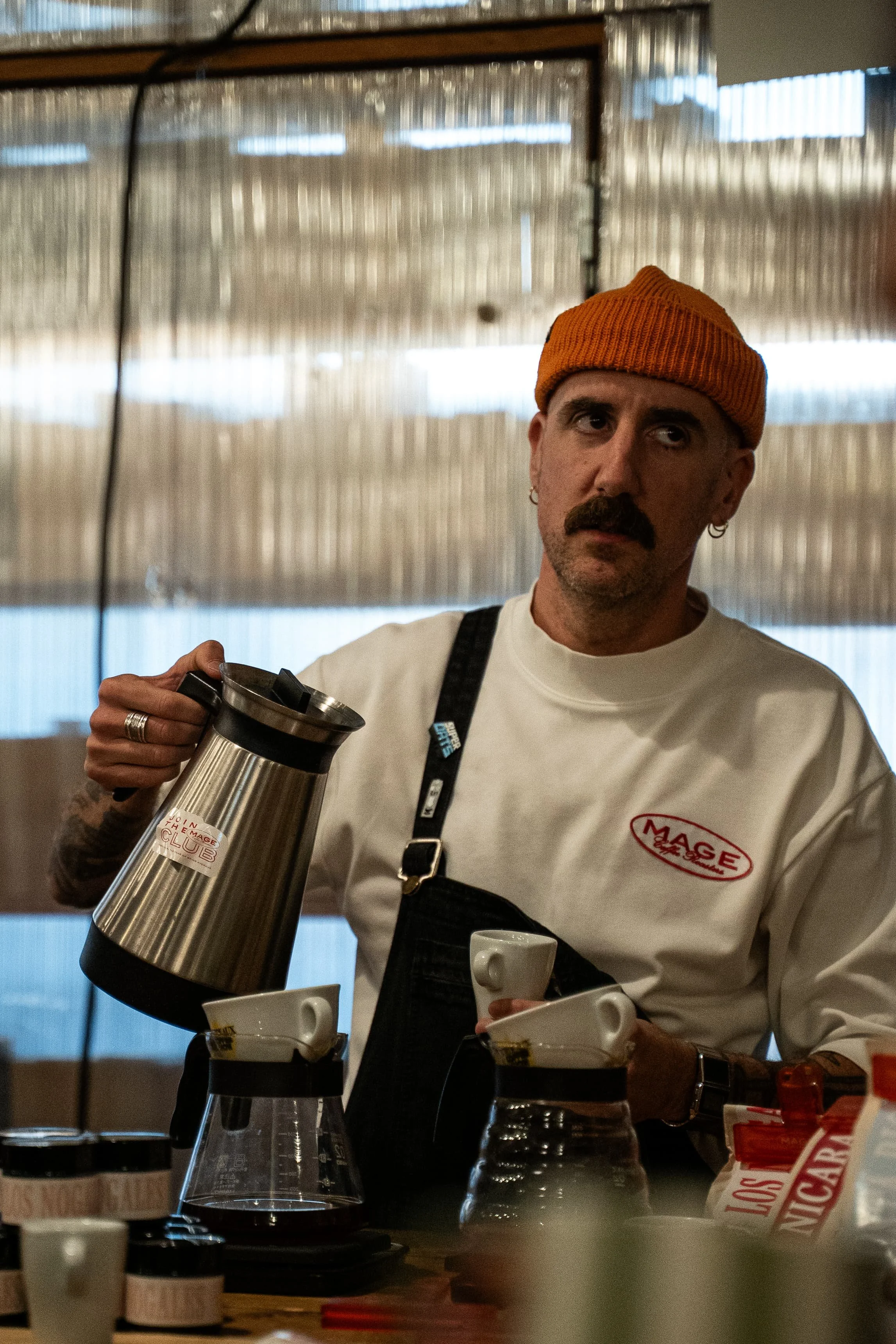 Homme avec un bonnet orange, moustache et boucles d'oreilles, tenant une cafetière en acier. Il porte un t-shirt blanc avec un logo rouge. Il se trouve dans un café, entouré de tasses et de cafetières, en train de préparer du café.