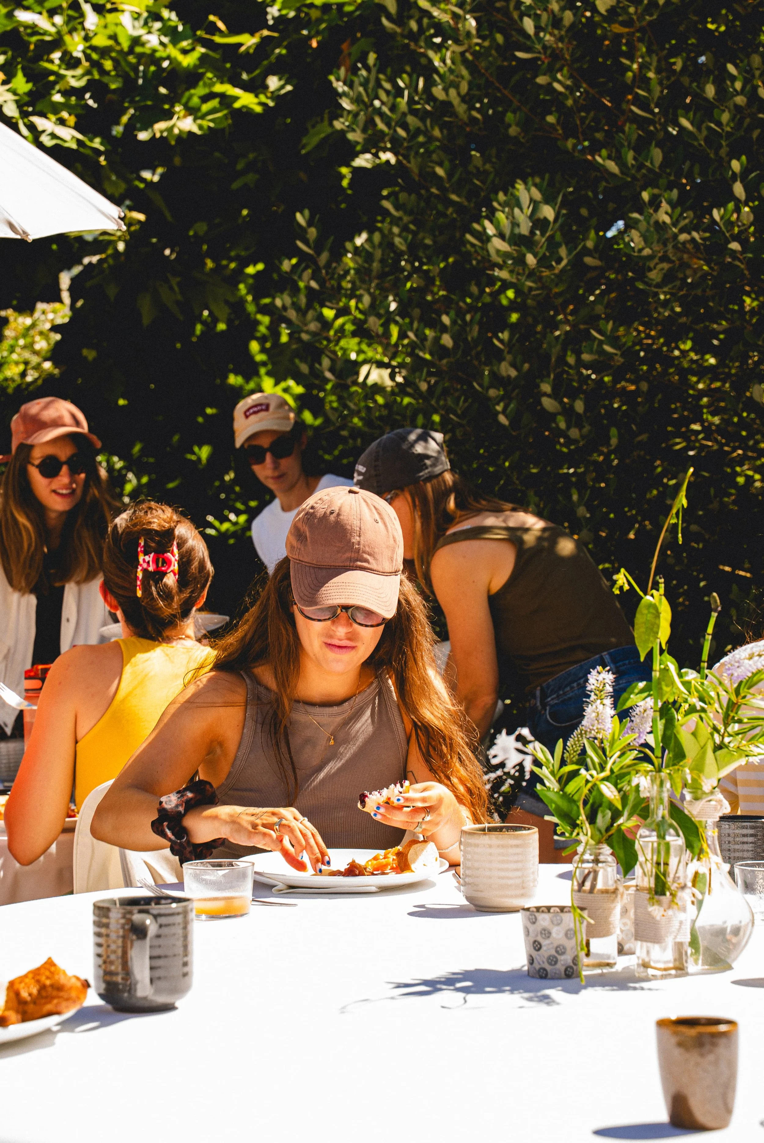 Un groupe de personnes assises autour d'une table lors d'un repas en plein air, avec des fleurs et des plantes en décor, sous un ciel ensoleillé.