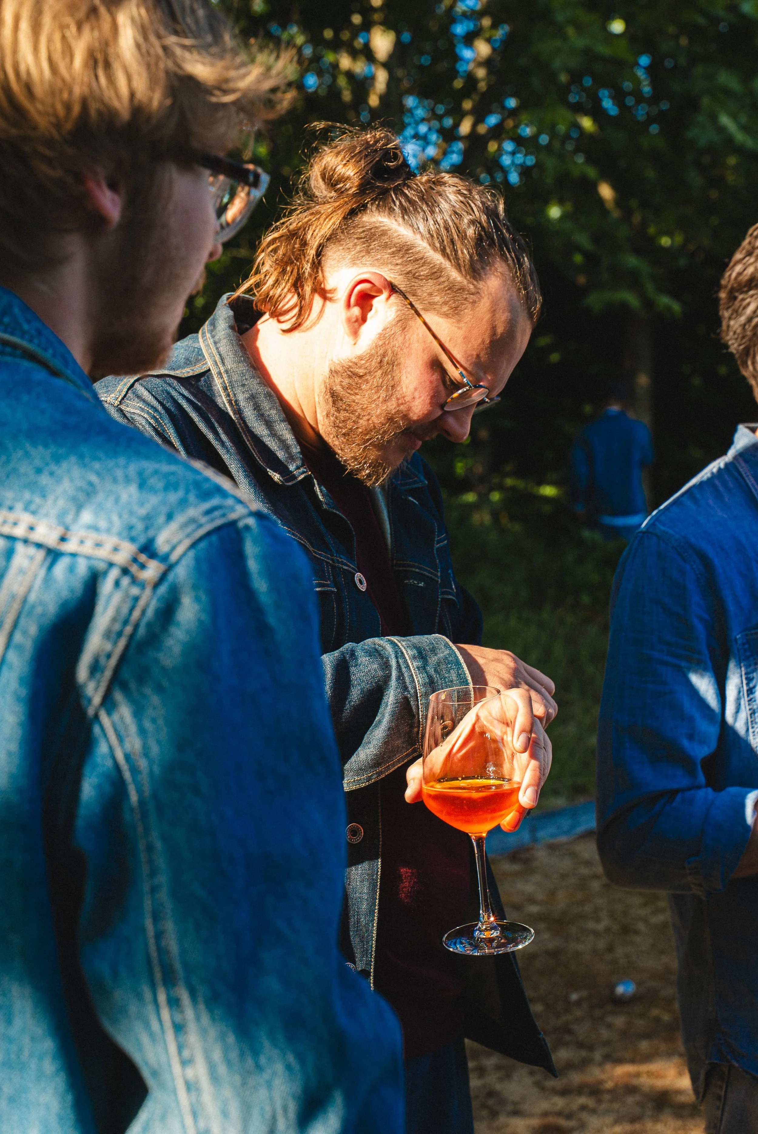 Trois personnes debout en extérieur, dont un homme tenant un verre de vin orange, le sourire et l'air détendu, probablement lors d'une dégustation ou d'un événement social parmi des arbres