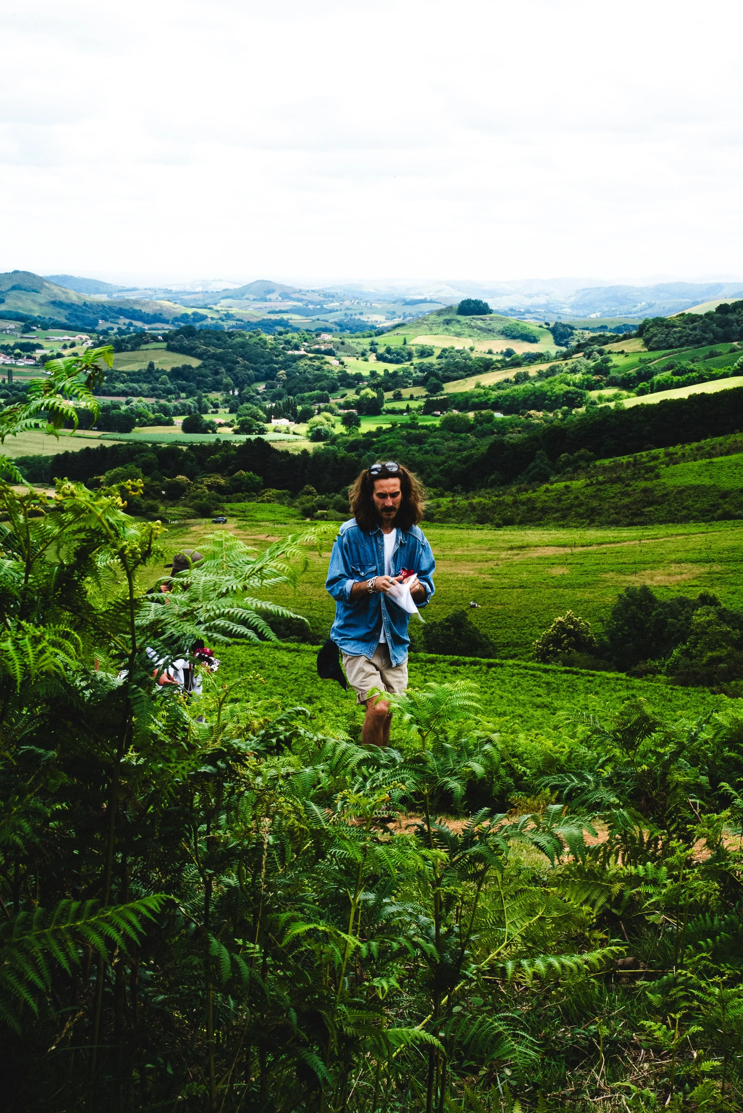Un homme avec des cheveux longs et une barbe marche dans une ferme entourée de verdure, avec un paysage vallonné au loin, sous un ciel nuageux.