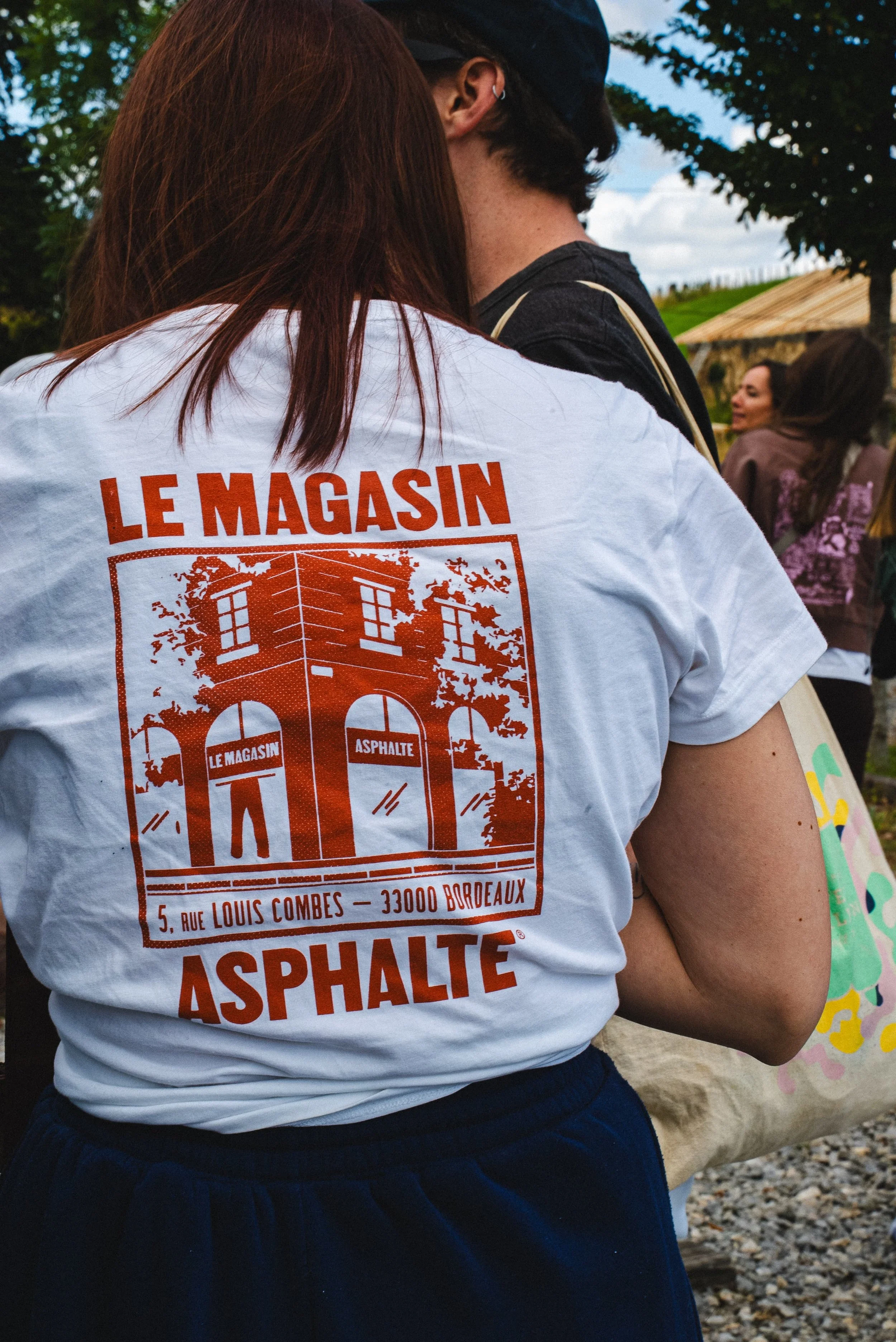 Une femme avec des cheveux bruns porte un t-shirt blanc avec des inscriptions en rouge. Elle est près d'autres personnes dans un environnement extérieur avec des arbres et un ciel nuageux.