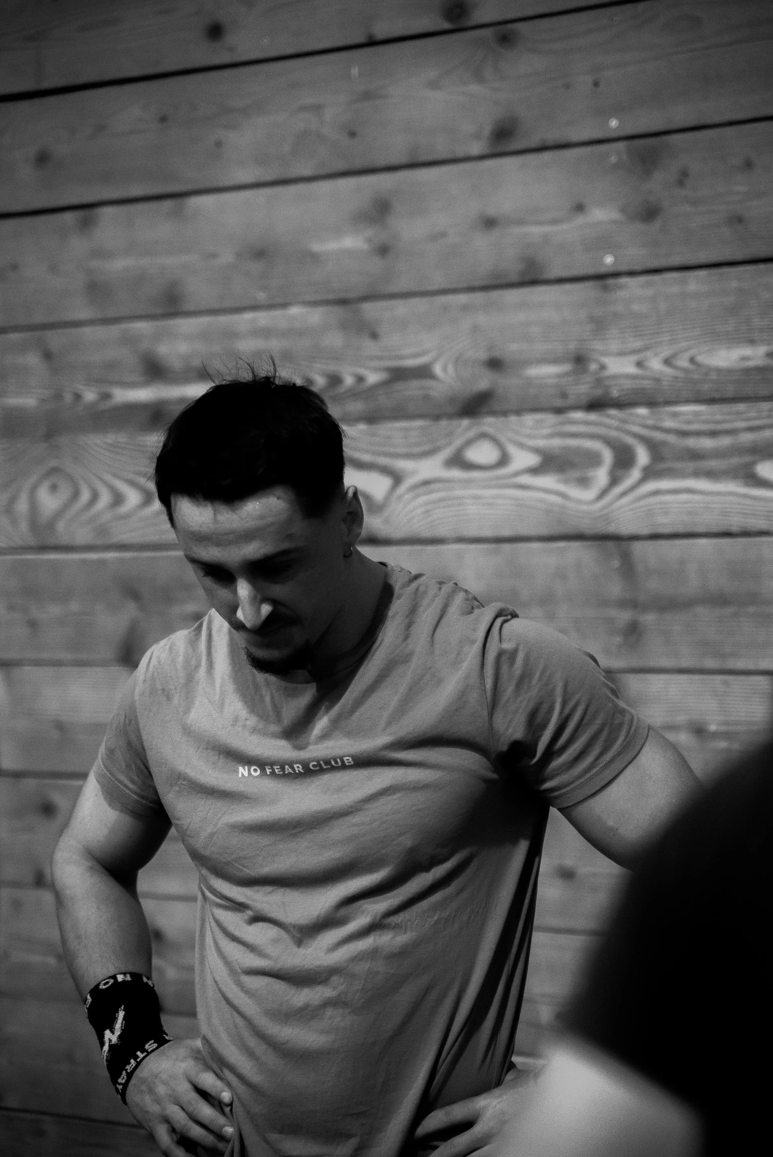 Une personne portant un t-shirt gris avec l'inscription "NO FEAR CLUB" et un bracelet noir, pose contre un mur en bois.