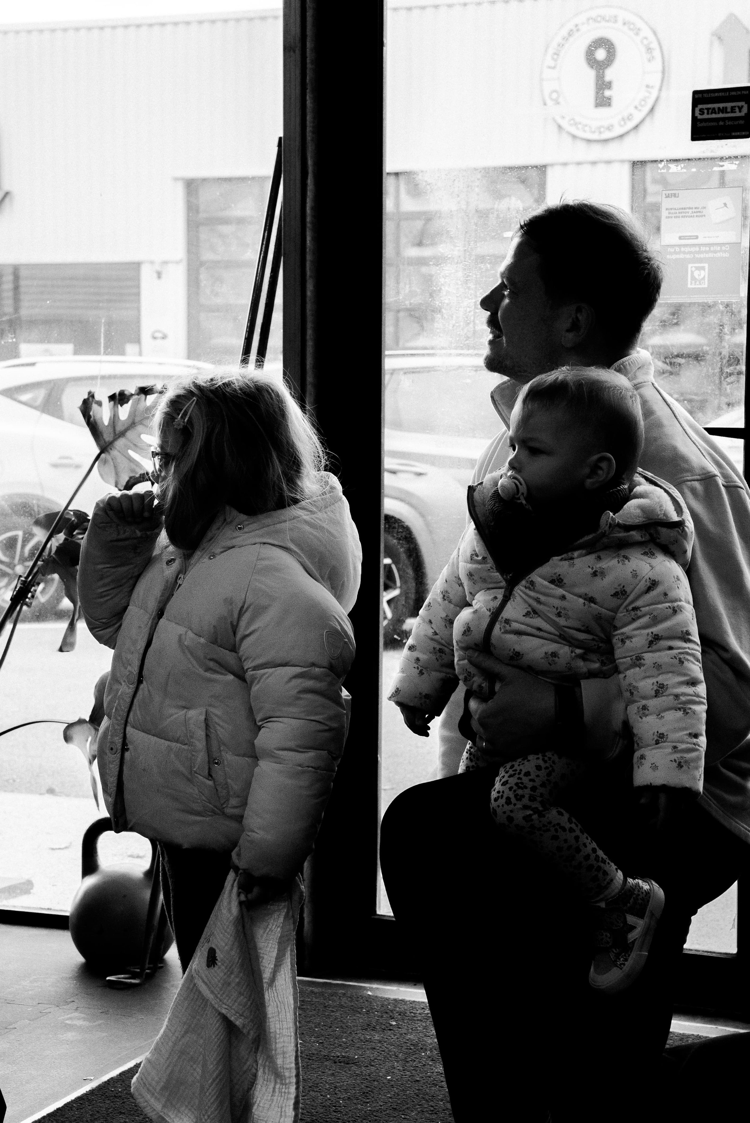 Une famille, composée d'un homme et de deux enfants, assise près d'une fenêtre dans une salle d'attente. La scène est en noir et blanc.