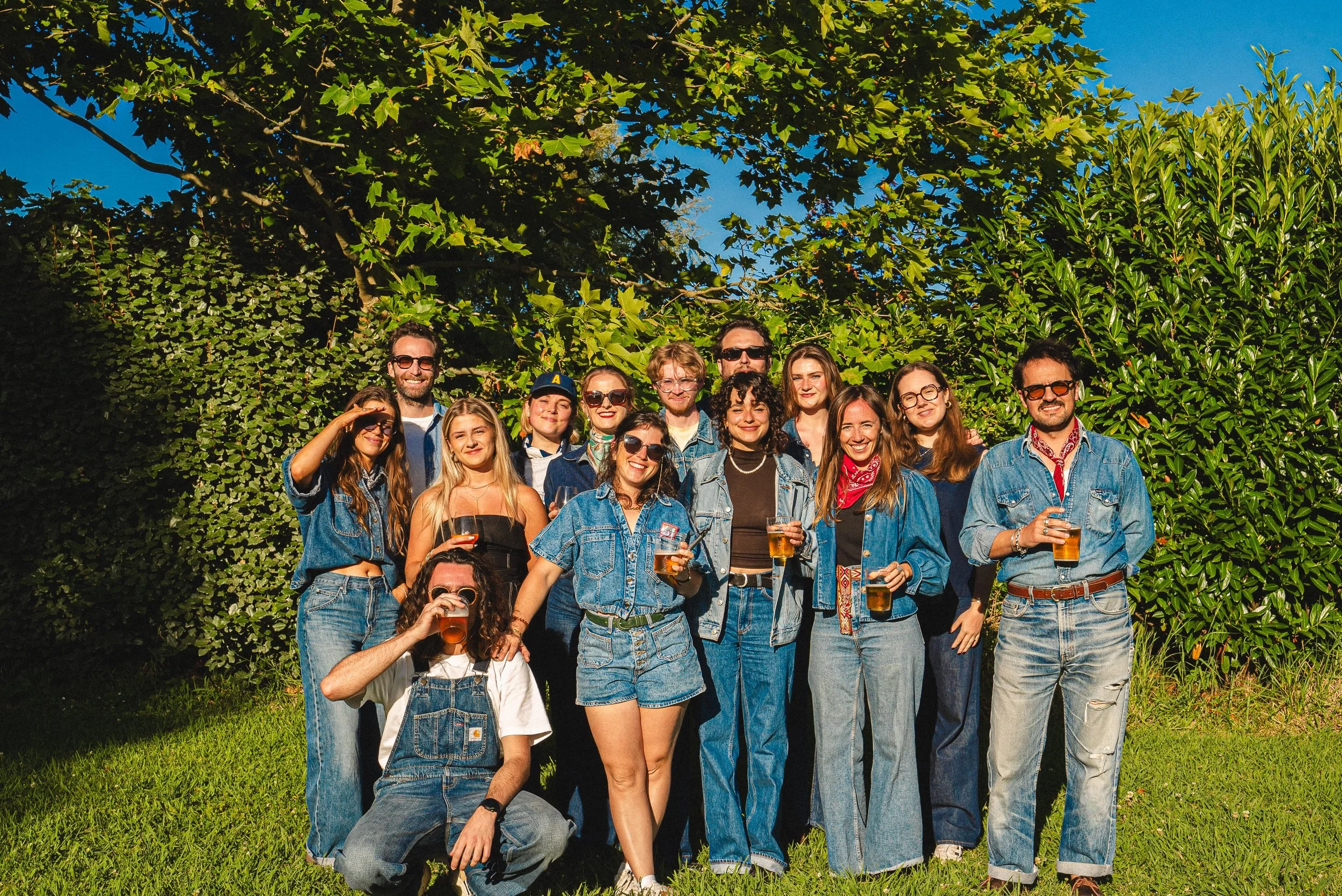 Groupe de personnes souriantes posant à l'extérieur, portant des vêtements en jean, certains avec des lunettes de soleil, tenant des verres de bière, sous un ciel bleu ensoleillé avec des arbres à feuilles vertes en arrière-plan.
