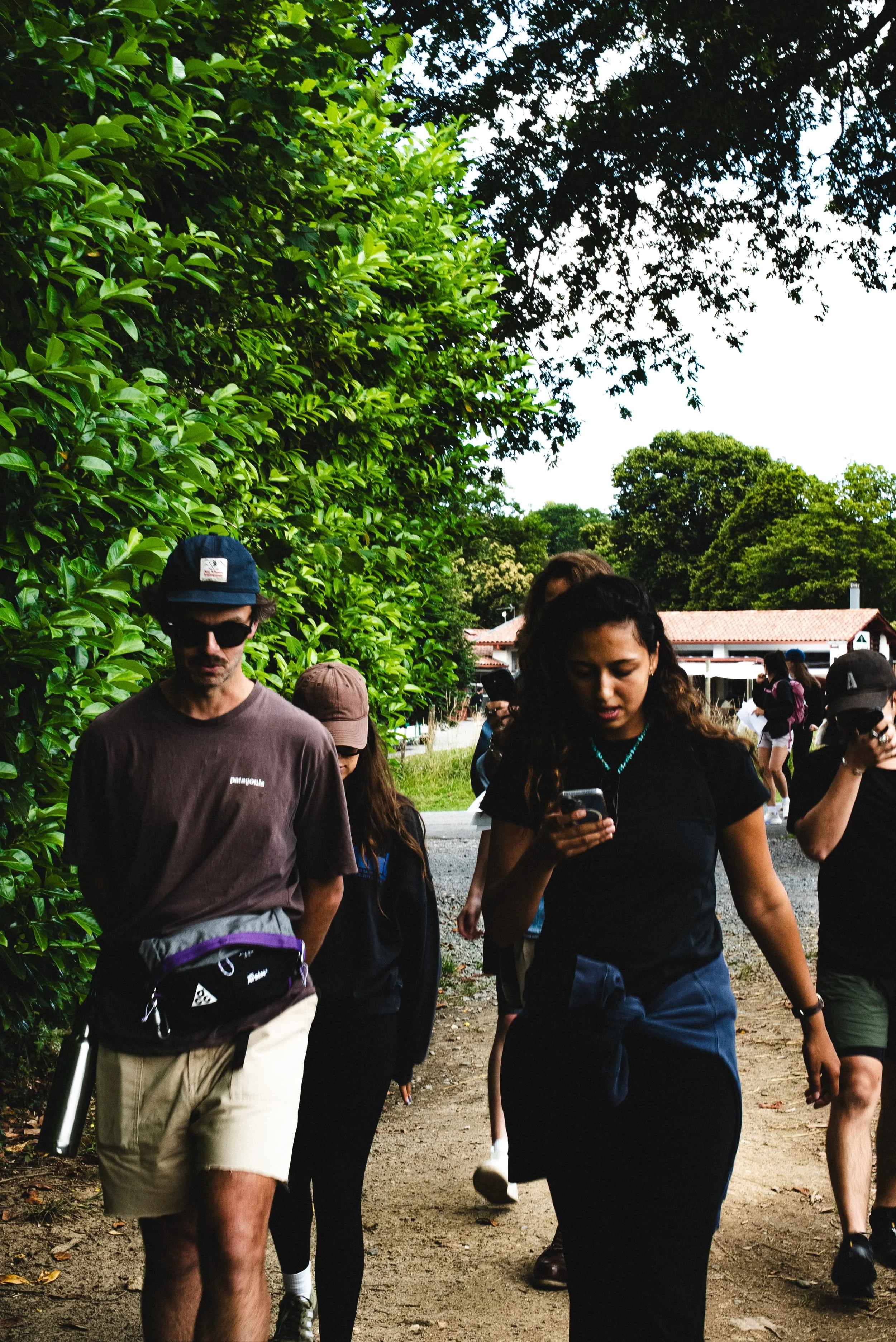 Groupe de personnes marchant dans un parc ou un environnement extérieur vert, avec des arbres et des bâtiments en arrière-plan, certains utilisant leurs téléphones portables.