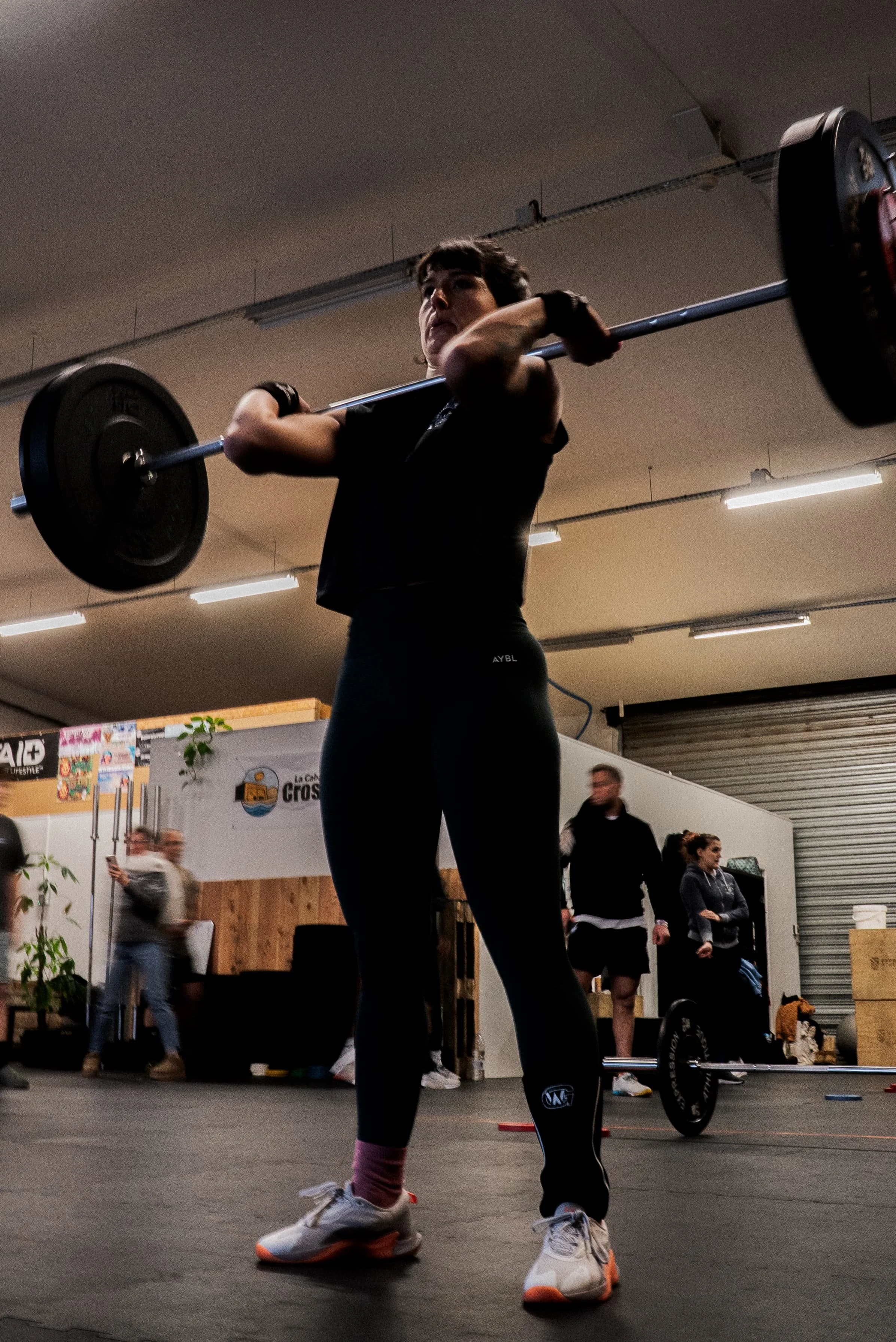 Une femme en sport soulève une barre avec poids dans une salle de sport, entourée de plusieurs personnes observant.