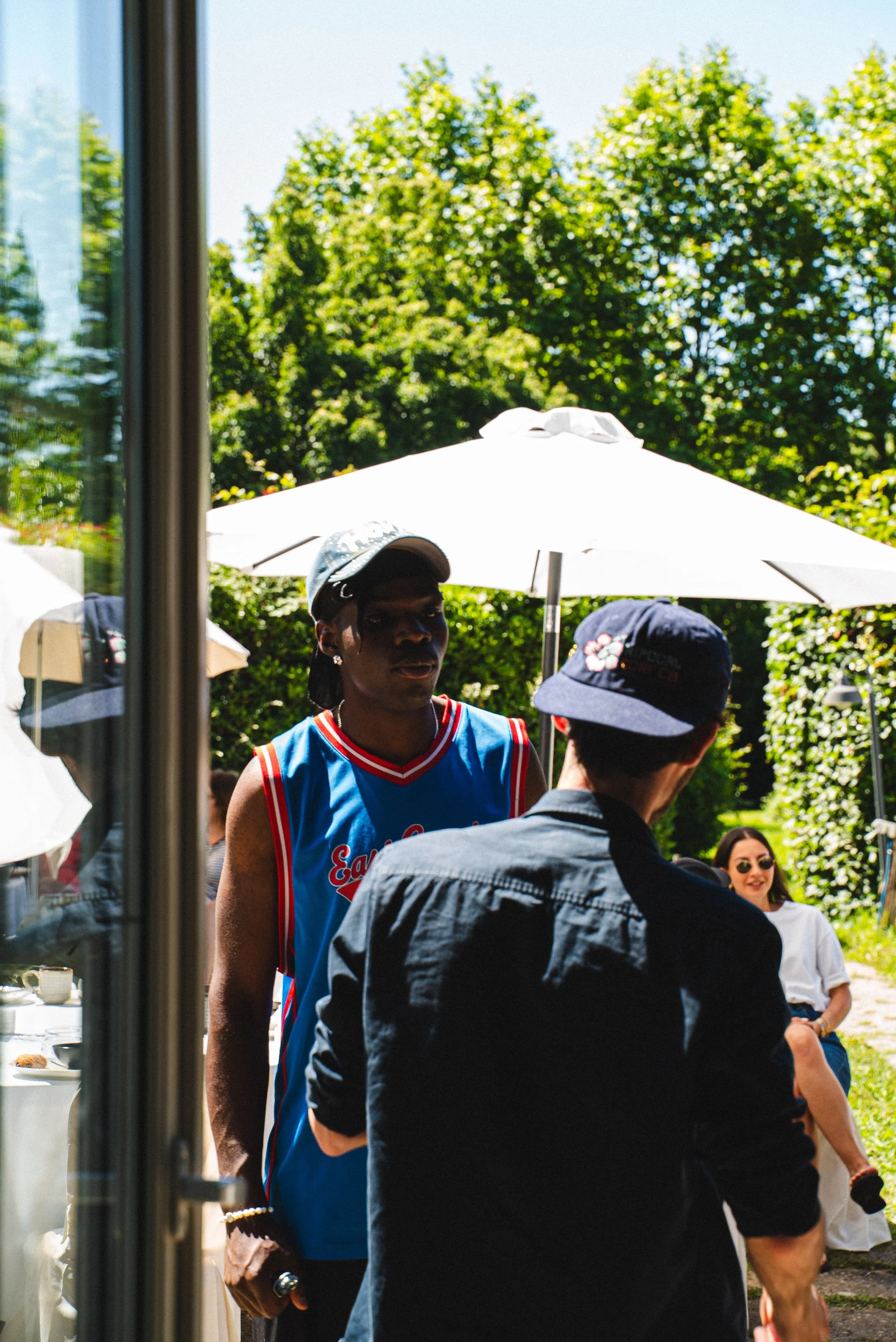 Une femme portant un maillot de basket bleu avec des détails rouges et blancs, portant une casquette grise, en train de parler avec un homme portant un chapeau noir et un t-shirt noir, lors d'un événement en plein air sous un parasol blanc, avec d'au