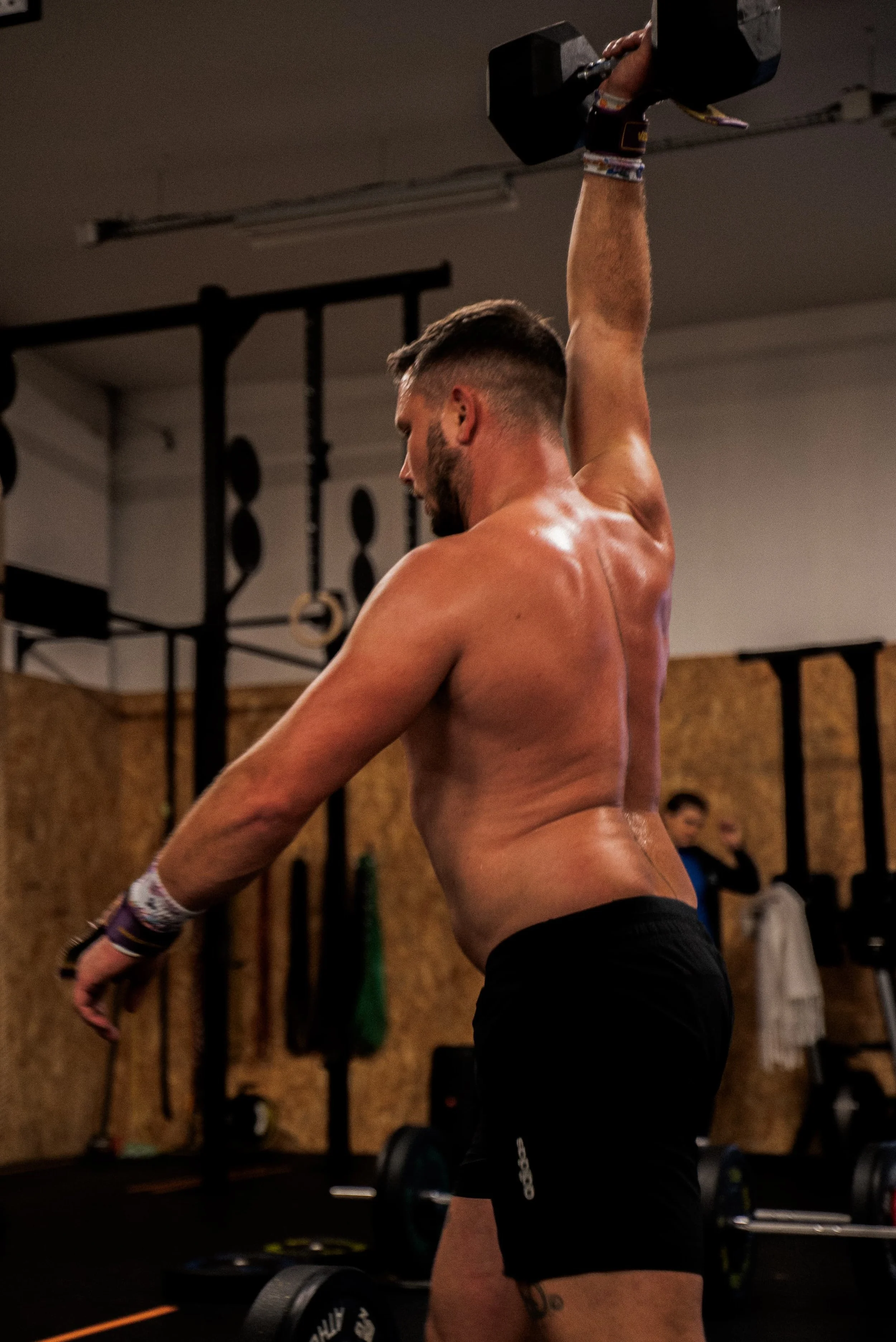 Un homme nain dans une salle de sport, portant un bracelet en caoutchouc, tenant un haltère au-dessus de sa tête, avec des poids et du matériel d'entraînement en arrière-plan.