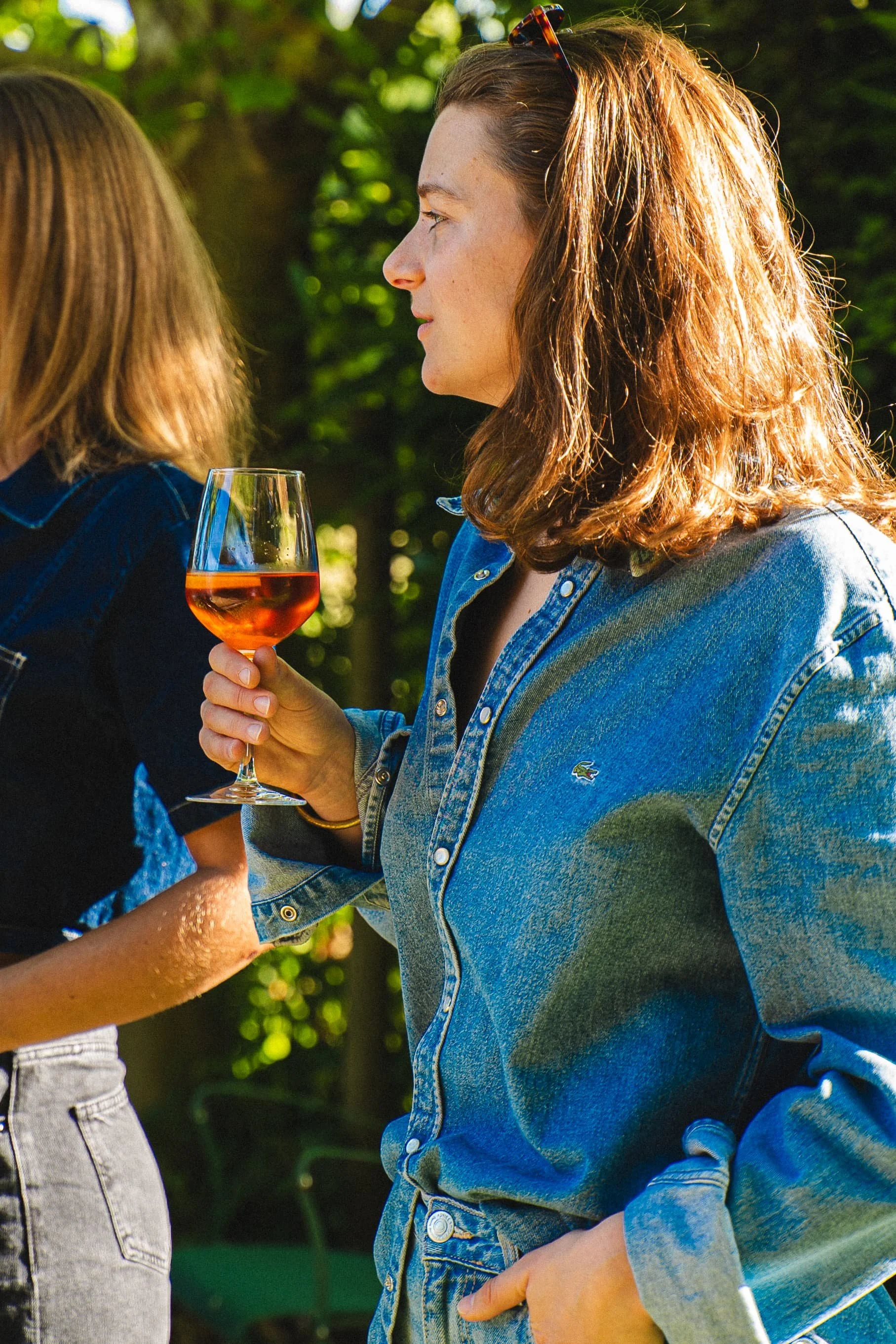 Une jeune femme tient un verre de vin rosé lors d'une rencontre en plein air, vêtue d'une veste en jean, dans un environnement greenery ensoleillé.