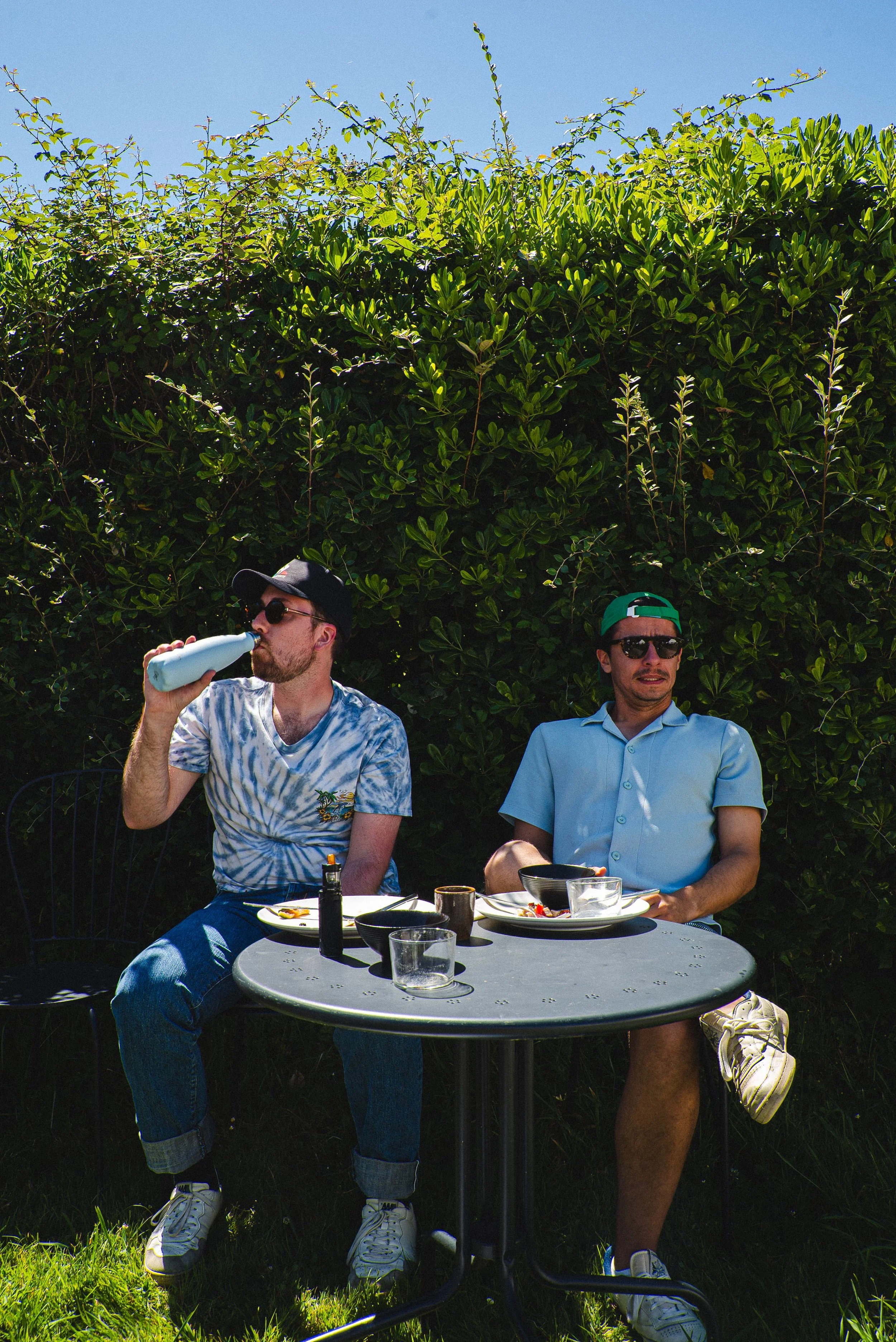 Deux hommes assis à une table de jardin en train de manger, l'un boit dans une bouteille, l'autre regarde avec une expression confuse, entourés de verdure et un ciel bleu.
