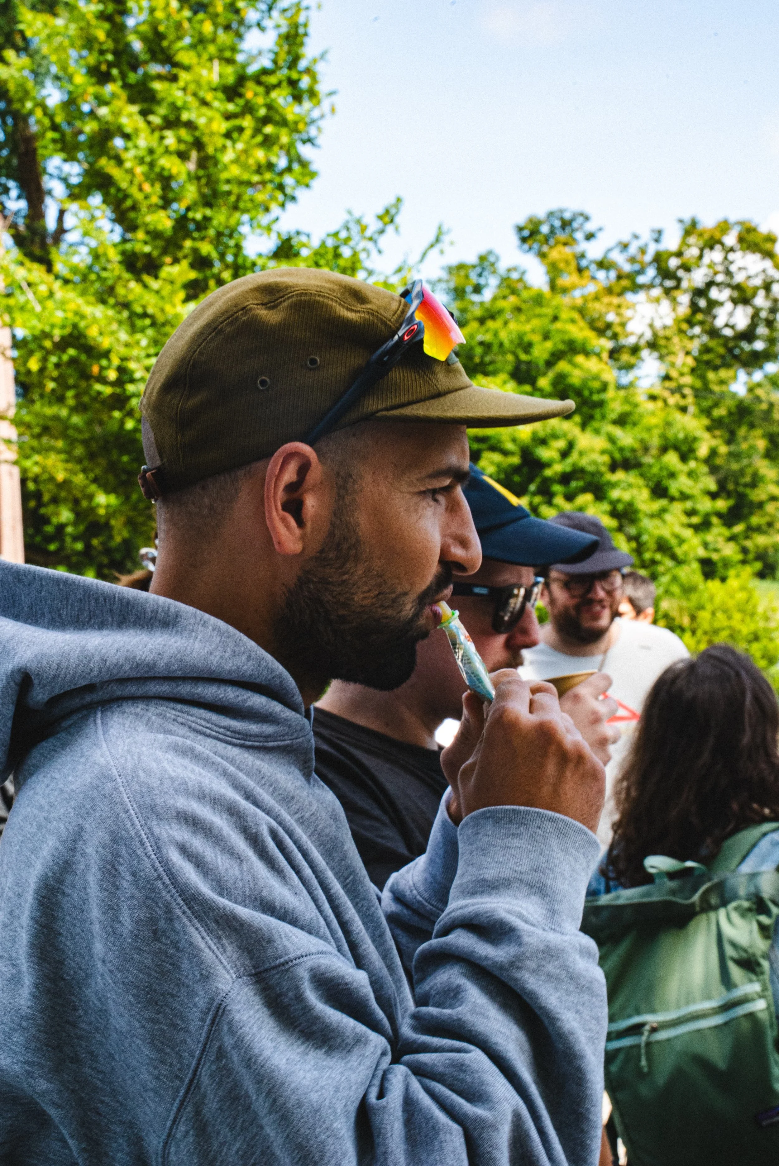 Groupe de personnes en extérieur, une personne en premier plan porte une casquette et des lunettes de soleil, elle tient un fruit ou un snack. Arrière-plan avec arbres verts.