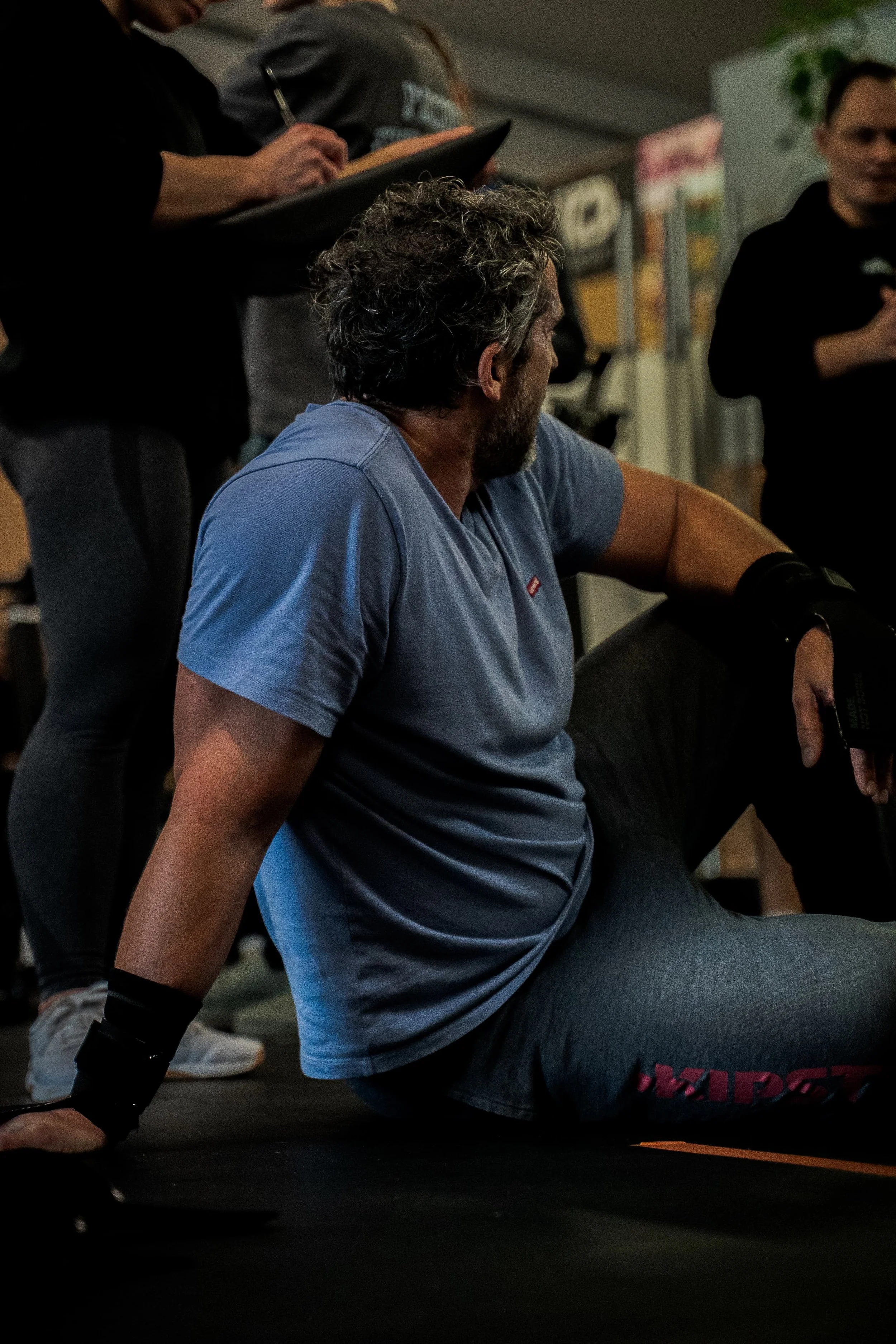 Une personne avec des cheveux bouclés gris et une barbe, portant un t-shirt bleu, réalise un exercice en position de pompe dans une salle, avec d'autres personnes en arrière-plan