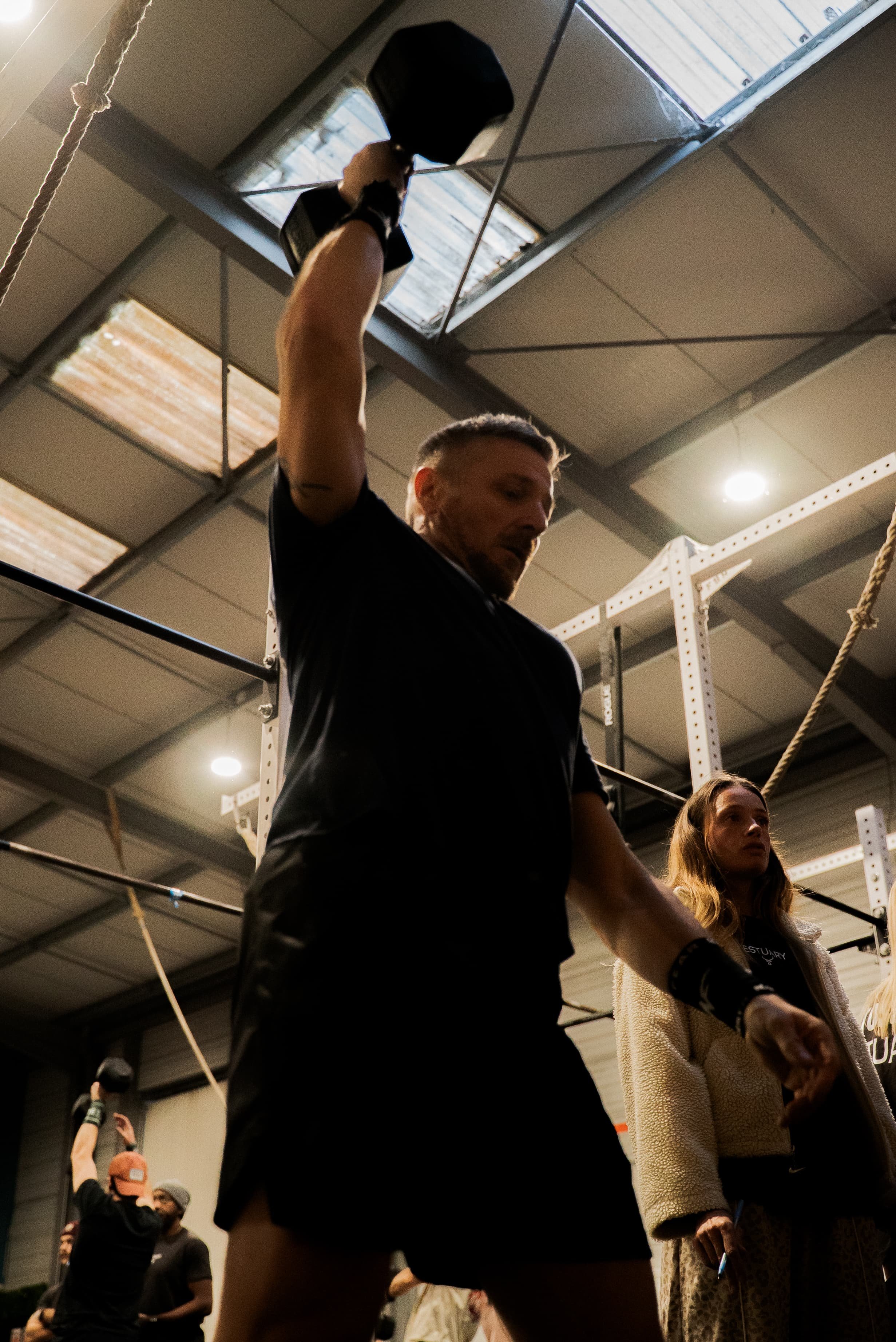 Groupe de personnes dans une salle de sport ou un gymnase, certaines tenant des haltères. Le plafond est fait de panneaux en métal et il y a des barres de traction. L'ambiance est sportive et décontractée.