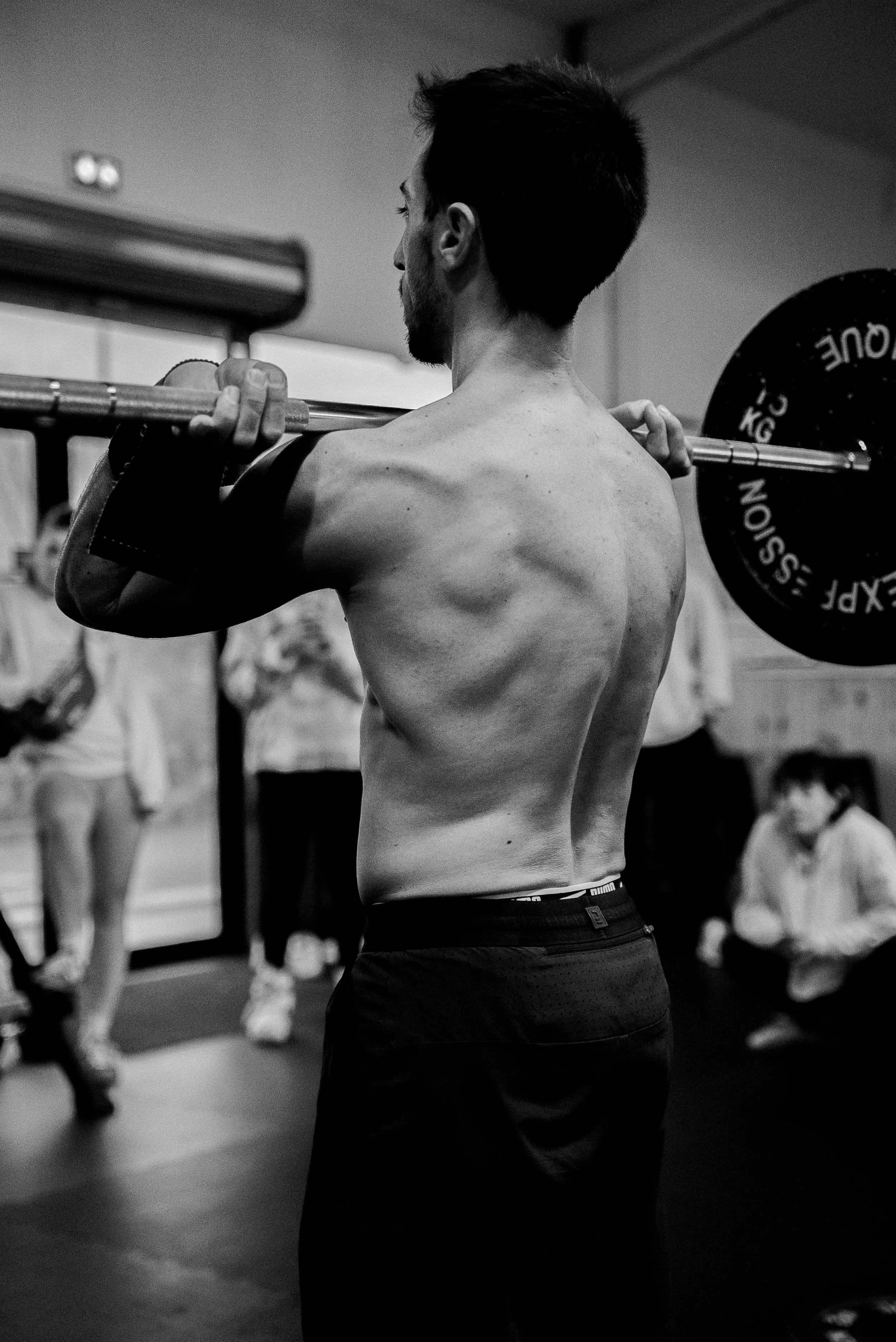 Un homme musclé, vu de dos, en train de faire un squat avec une barre chargée sur ses épaules dans une salle de sport.