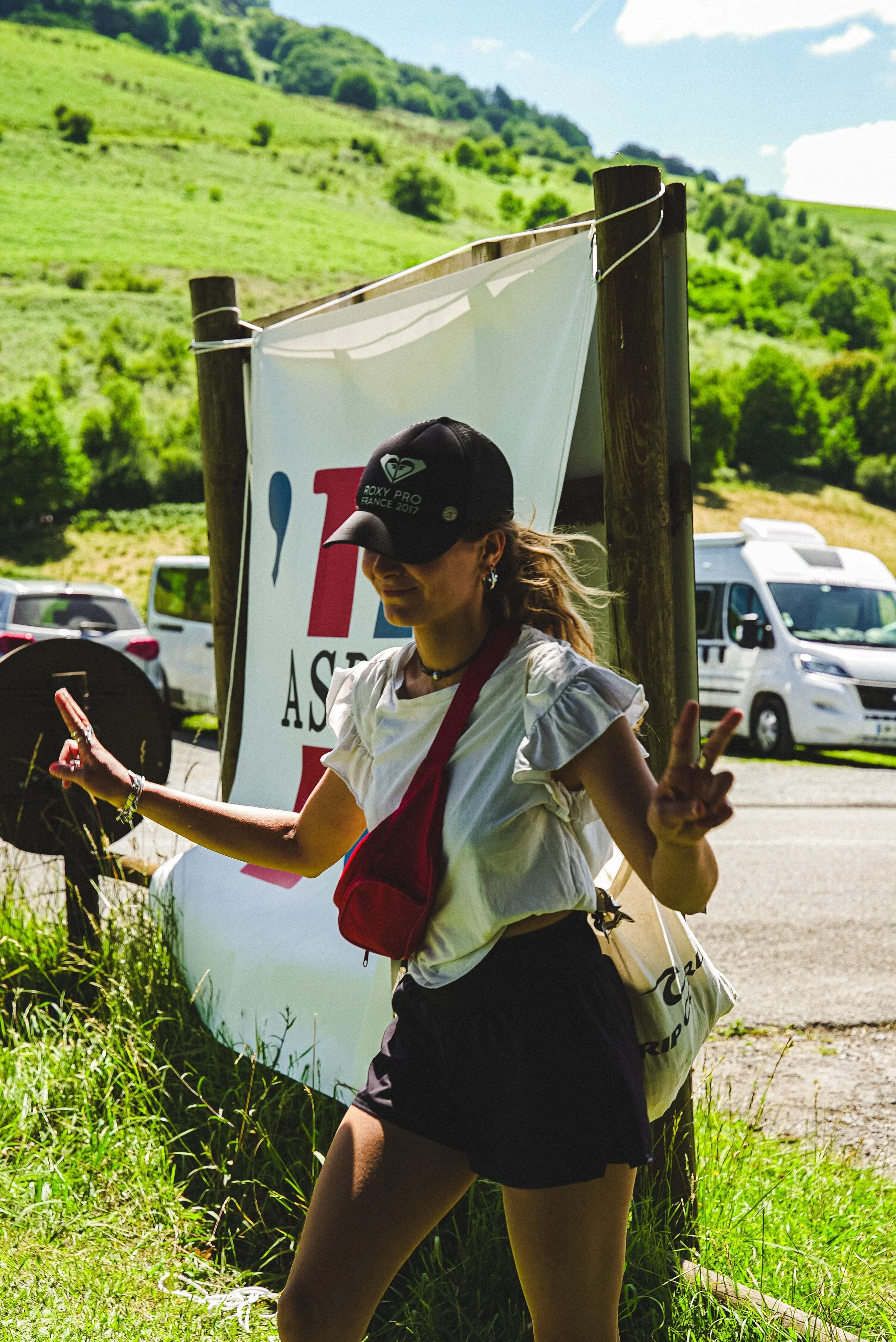 Jeune femme en tenue sportive, portant une casquette avec le logo Roxy, faisant un signe de paix avec ses doigts, en plein air avec un fond de nature verdoyante et un panneau avec le texte 'I ❤️ T'.