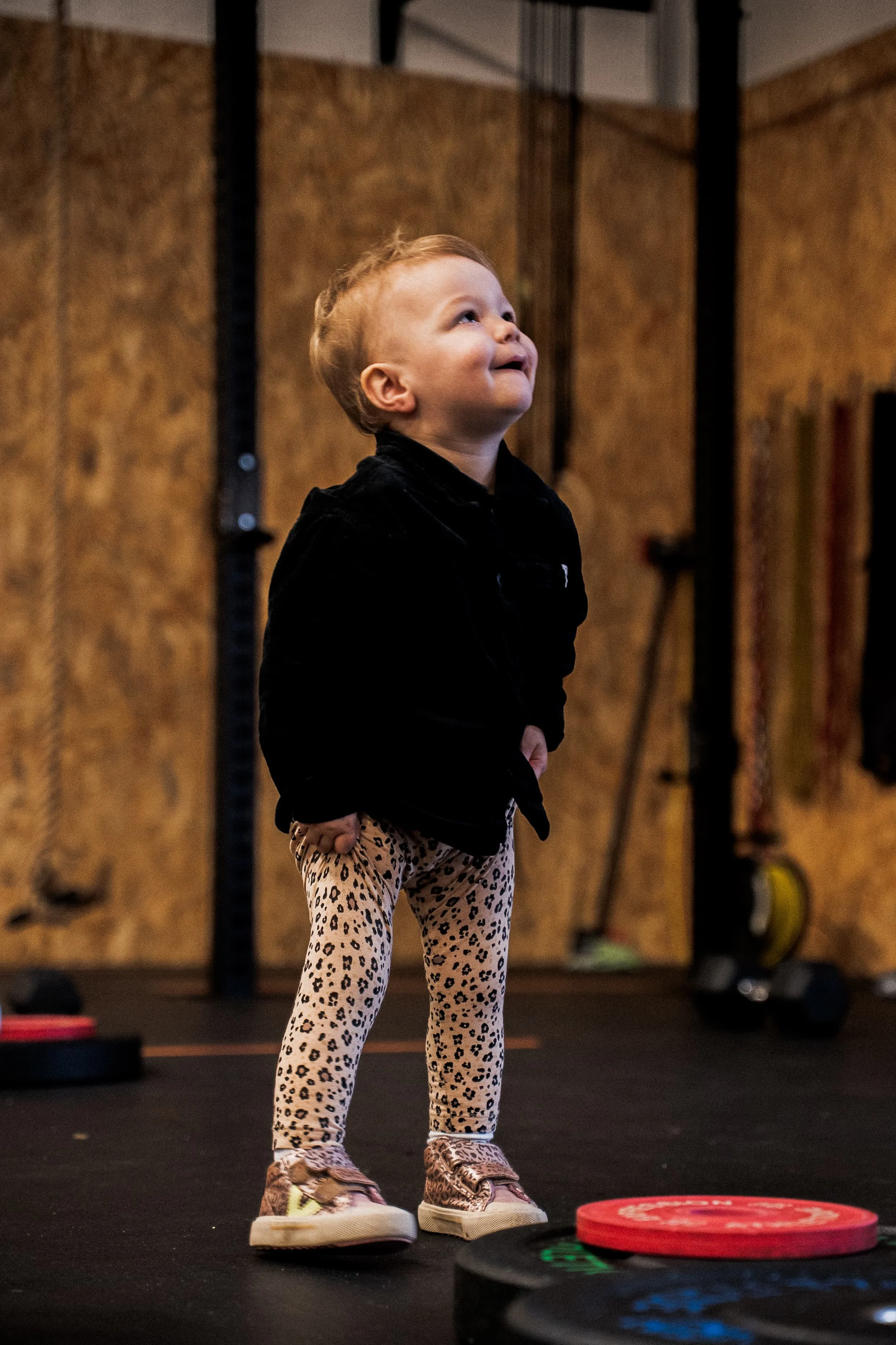Une jeune fille avec des cheveux courts, portant un haut noir et un pantalon à motifs léopard, debout dans une salle de sport ou dojo, regardant vers le haut avec un sourire, entourée d'équipements d'entraînement.