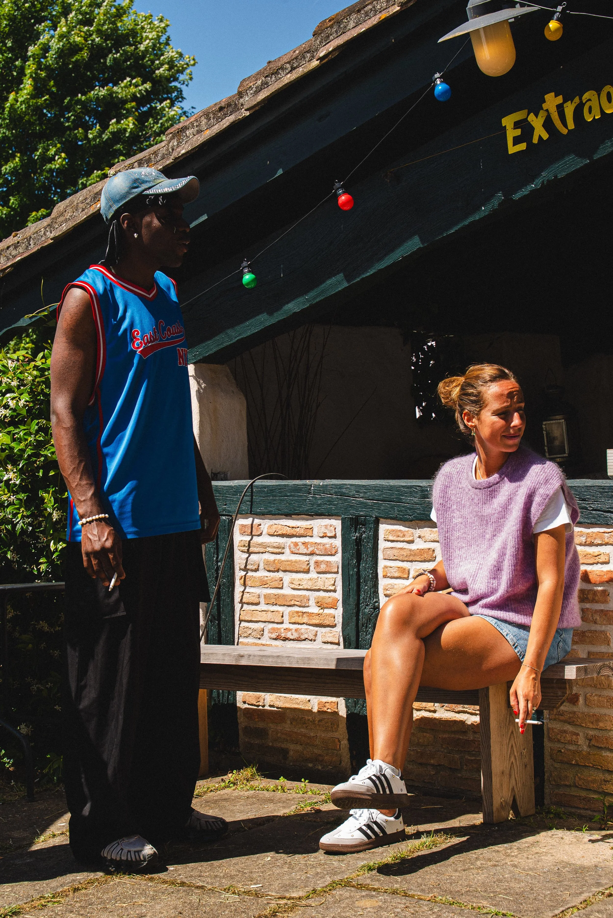 Une jeune femme assise sur un banc en bois, portant un pull violet et un short en denim, avec des baskets Adidas, semblant parler ou rire. À côté d'elle, un homme debout, portant une casquette, des lunettes de soleil, un maillot de sport bleu avec le