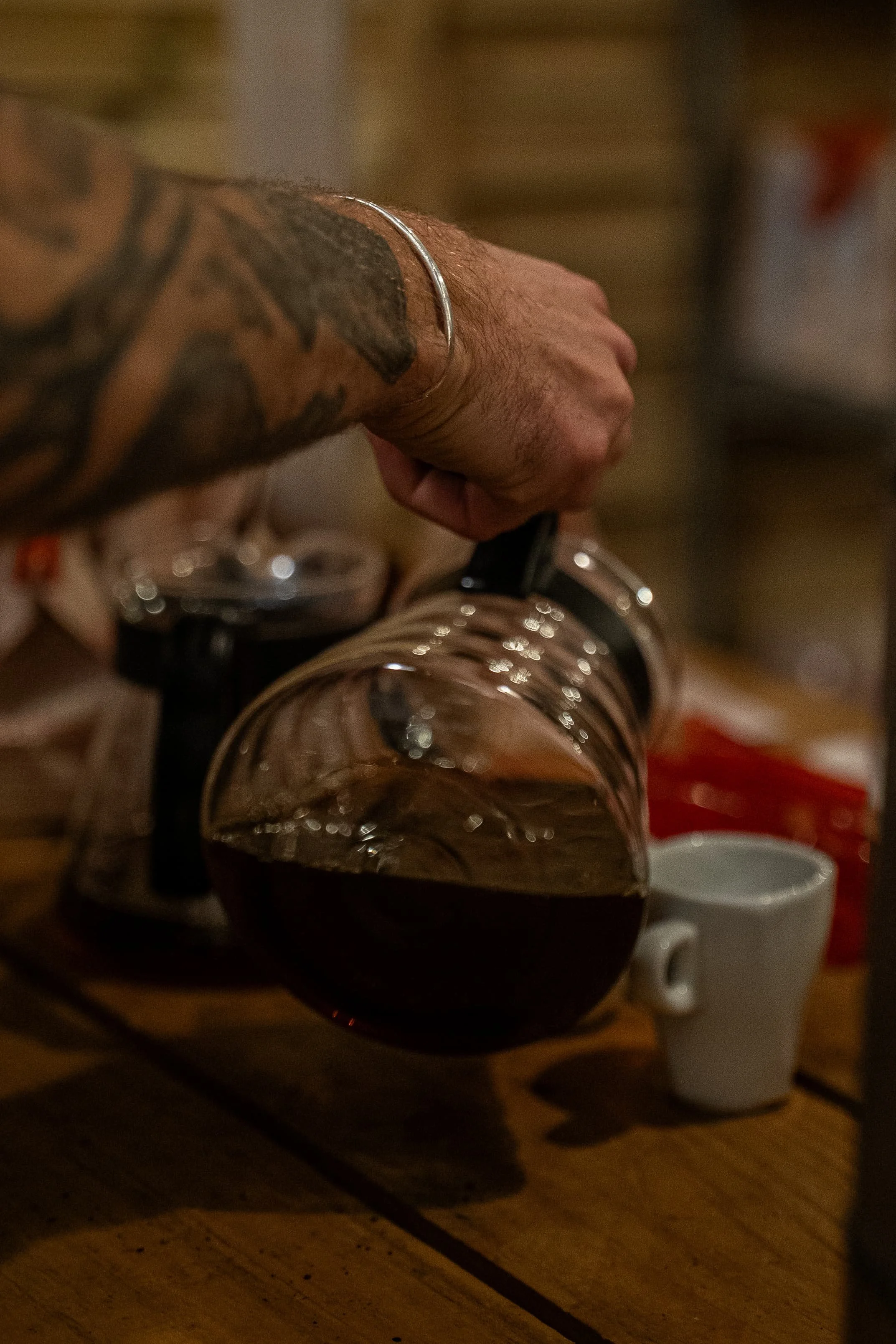 Une main tatouée tenant une tasse de café dans une cuisine, avec une théière et des tasses blanches en arrière-plan.