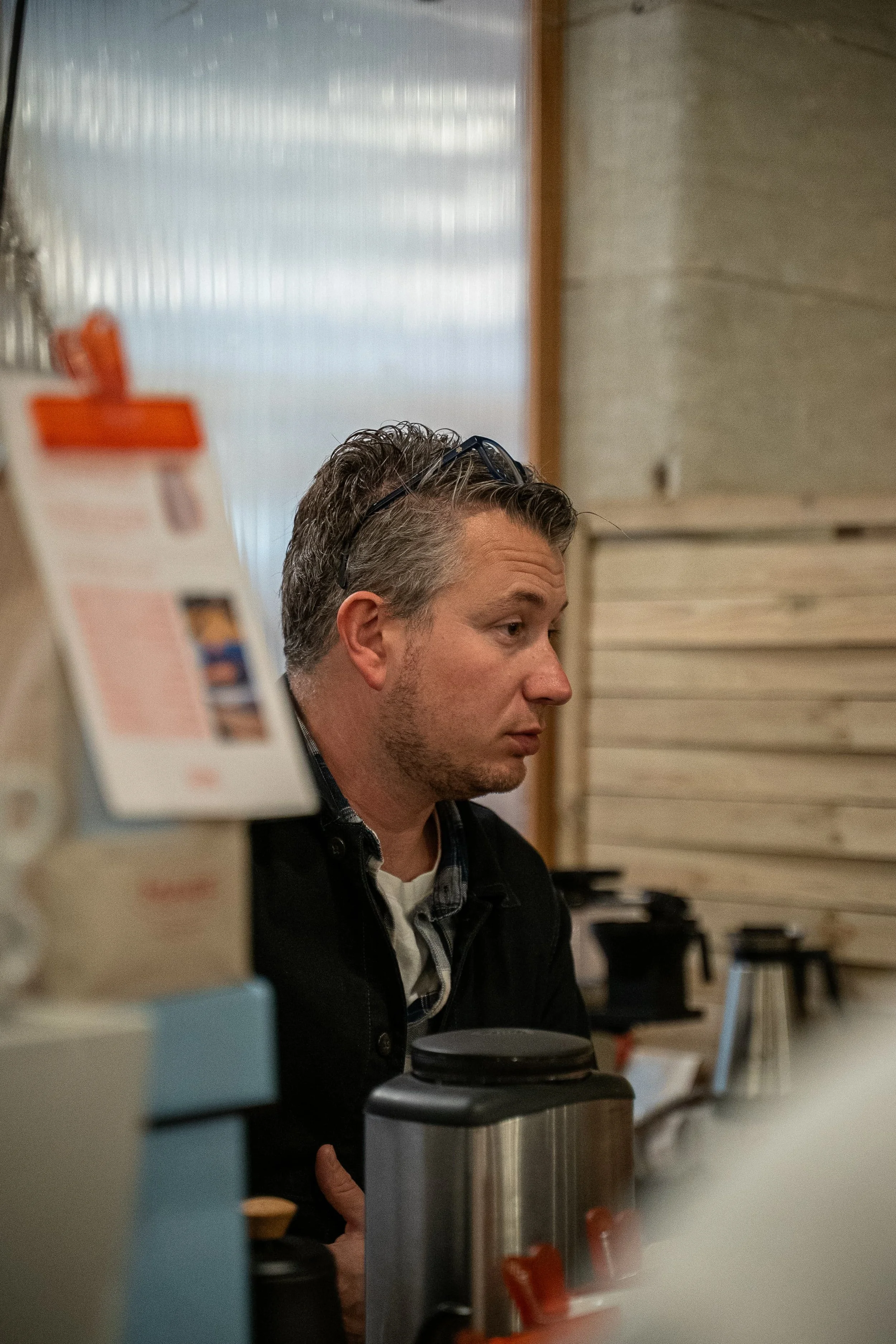 Un homme regarde vers la droite dans un environnement intérieur avec des murs en bois, entouré de bureau ou d'ustensiles, vu en profil.