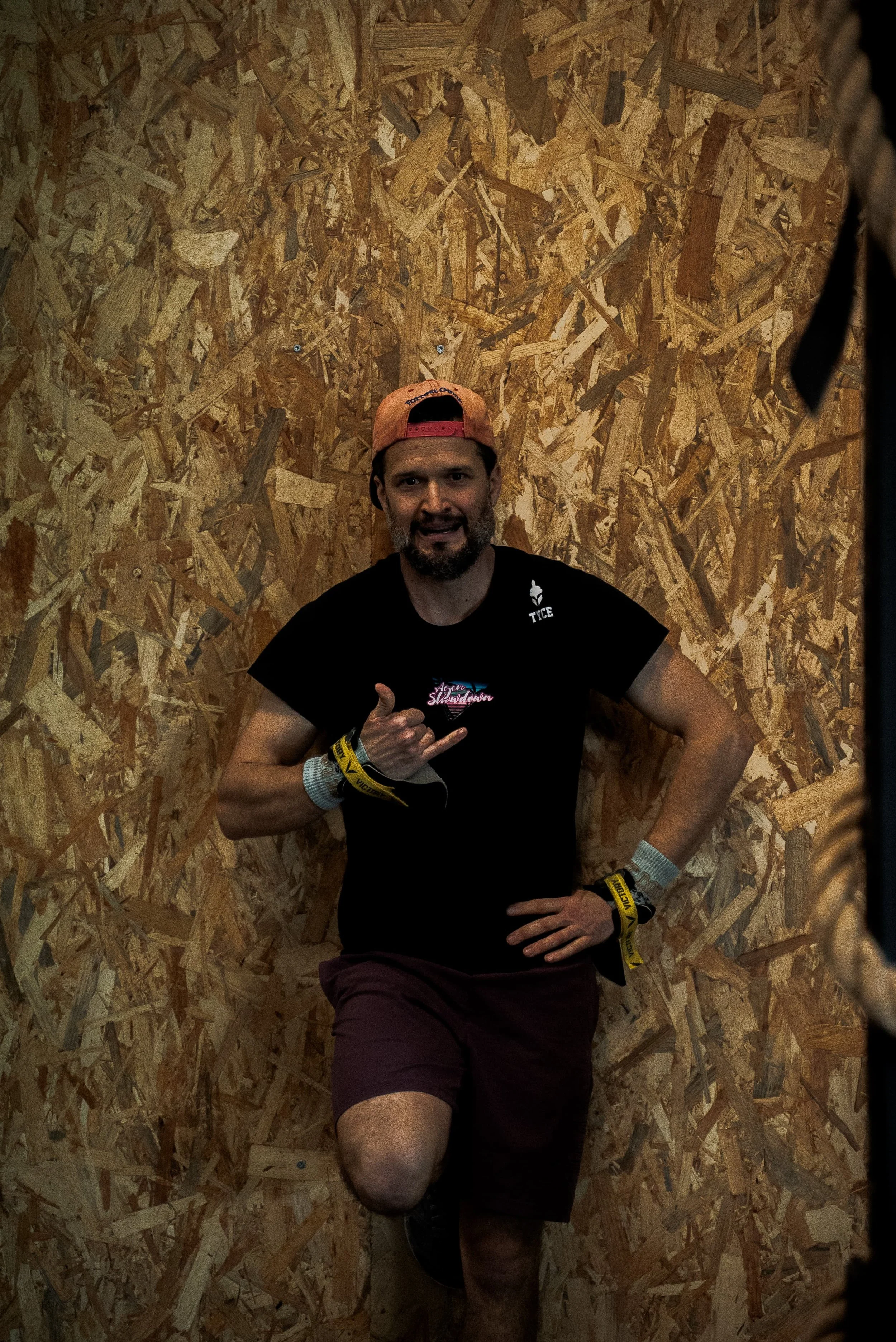 Un homme souriant portant un t-shirt noir, un bonnet orange à l'envers, et des bracelets, pose devant un mur en bois orné de morceaux de contreplaqué. Il fait un geste de la main avec un thumbs-up.