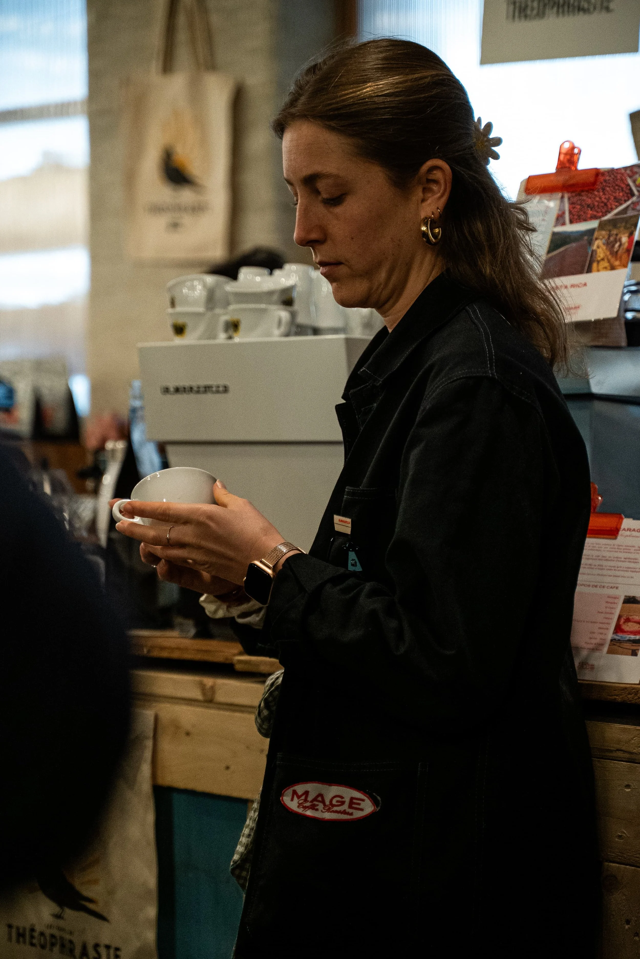 Femme portant un tablier noir avec un badge 'MAGE', tenant une tasse dans un café ou un restaurant.