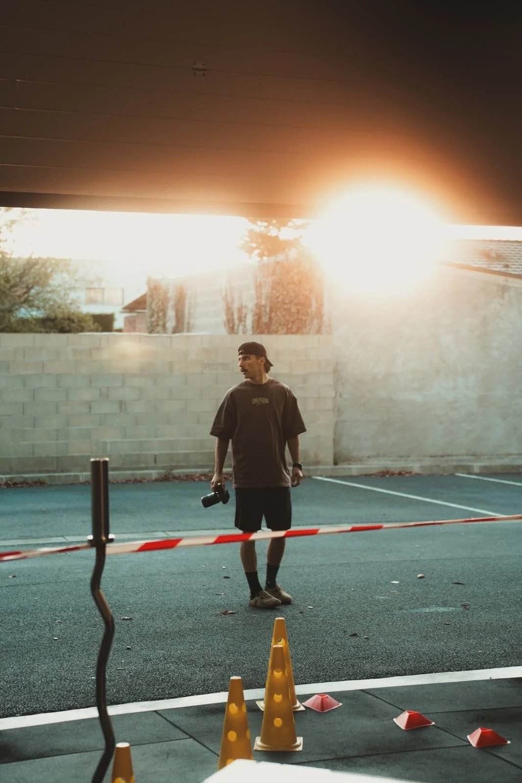 Un homme debout dans un parking en fin d'après-midi, tenant un appareil photo dans une main, avec un soleil couchant brillant derrière un mur en brique.