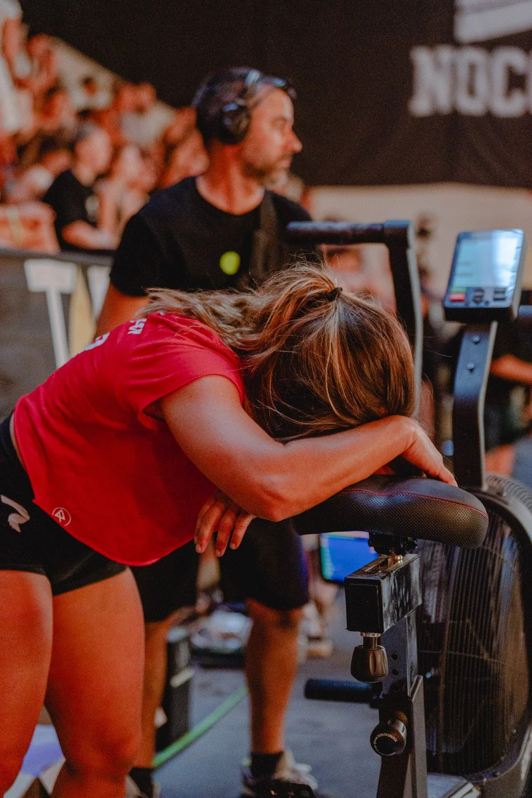 Femme épuisée en rouge s'appuyant sur un vélo stationnaire lors d'une compétition CrossFit — photographe CrossFit Voar by Tim