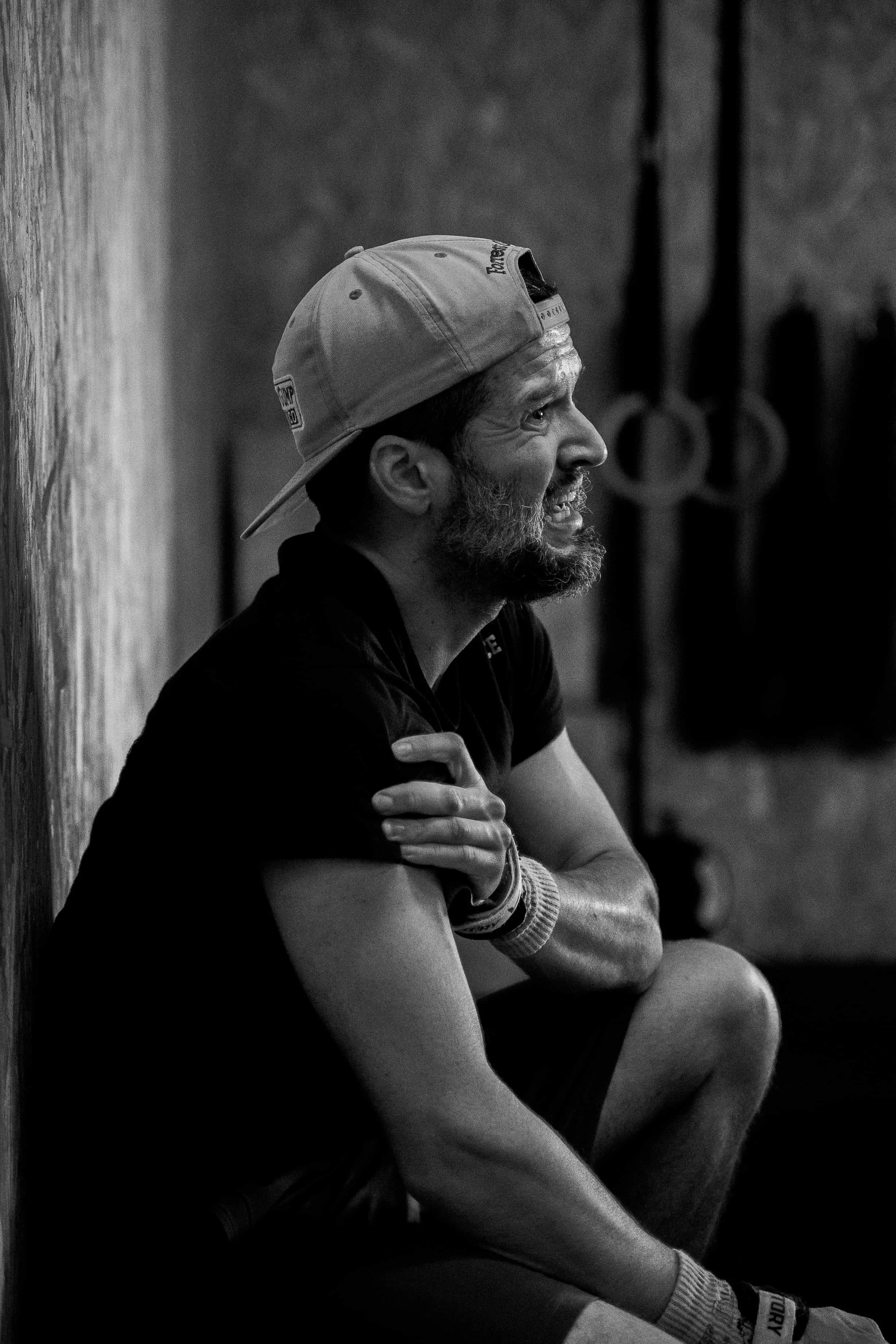 Un homme avec une barbe porte une casquette inclinée et un t-shirt noir, il est assis contre un mur en bois, souriant et regardant vers la droite.