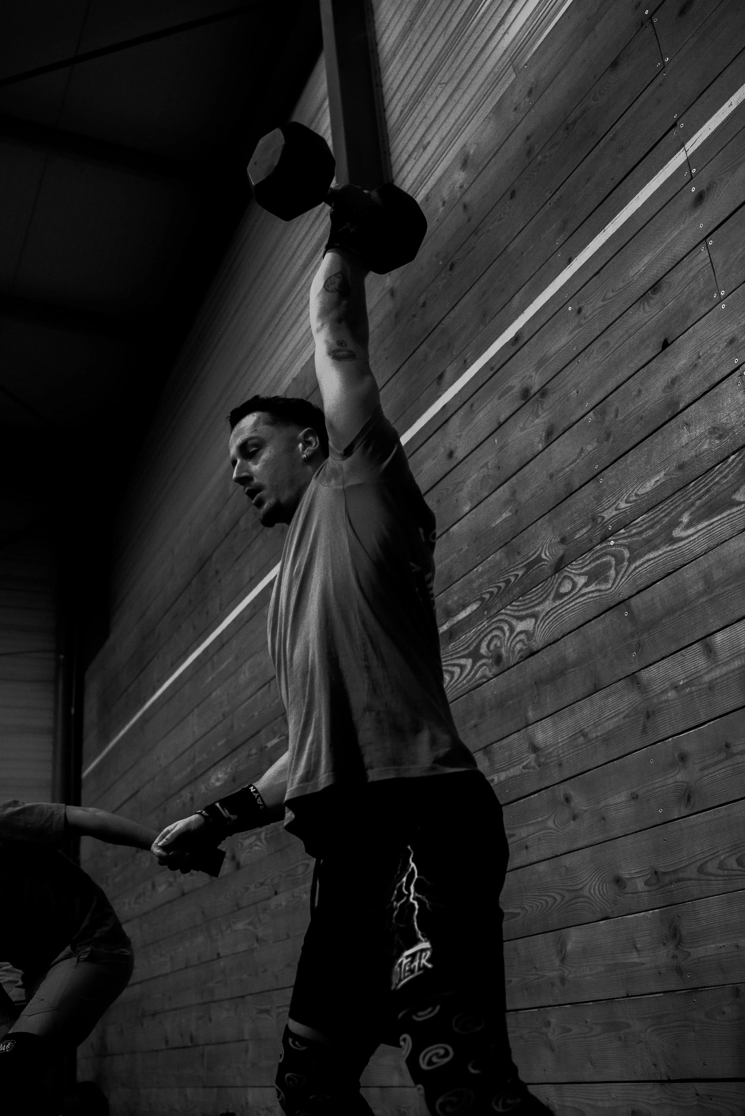 Un homme tenant un haltère en l'air dans une salle de sport avec un mur en bois en arrière-plan.