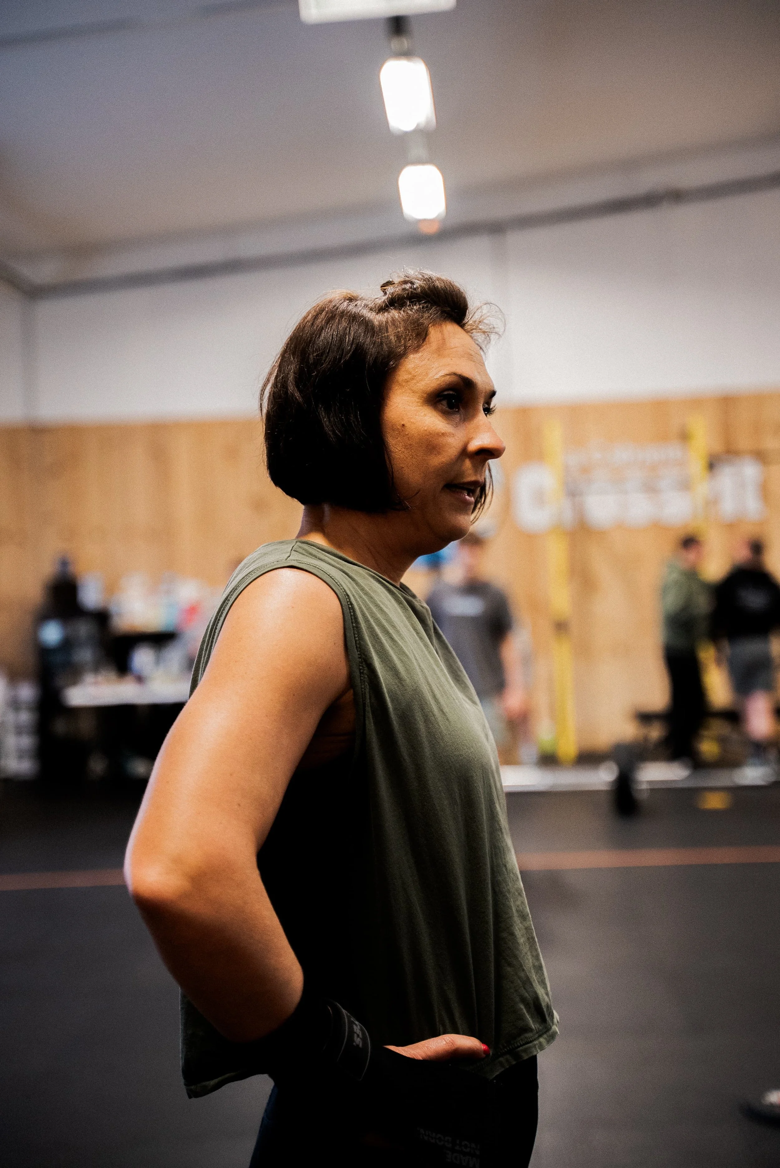 Femme avec une veste noire et un haut vert, debout dans une salle, probablement une salle d'entraînement ou un studio, portant un bracelet de poignet noir, avec un fond flou montrant d'autres personnes et des murs en bois.
