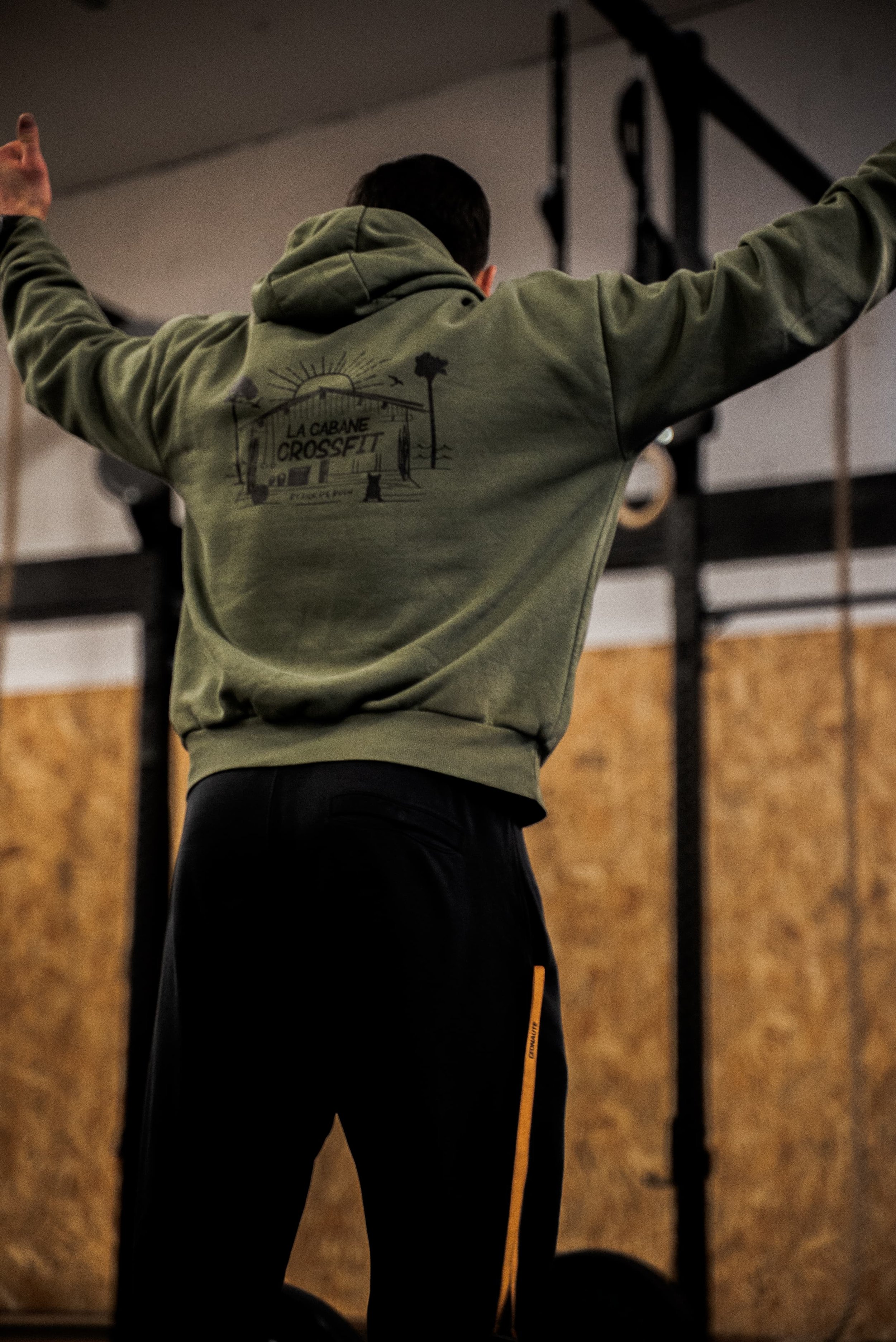 Homme vu de dos, portant un sweat à capuche vert avec un logo de cabane de crossfit, dans une salle de sport, avec des équipements d'entraînement en arrière-plan.