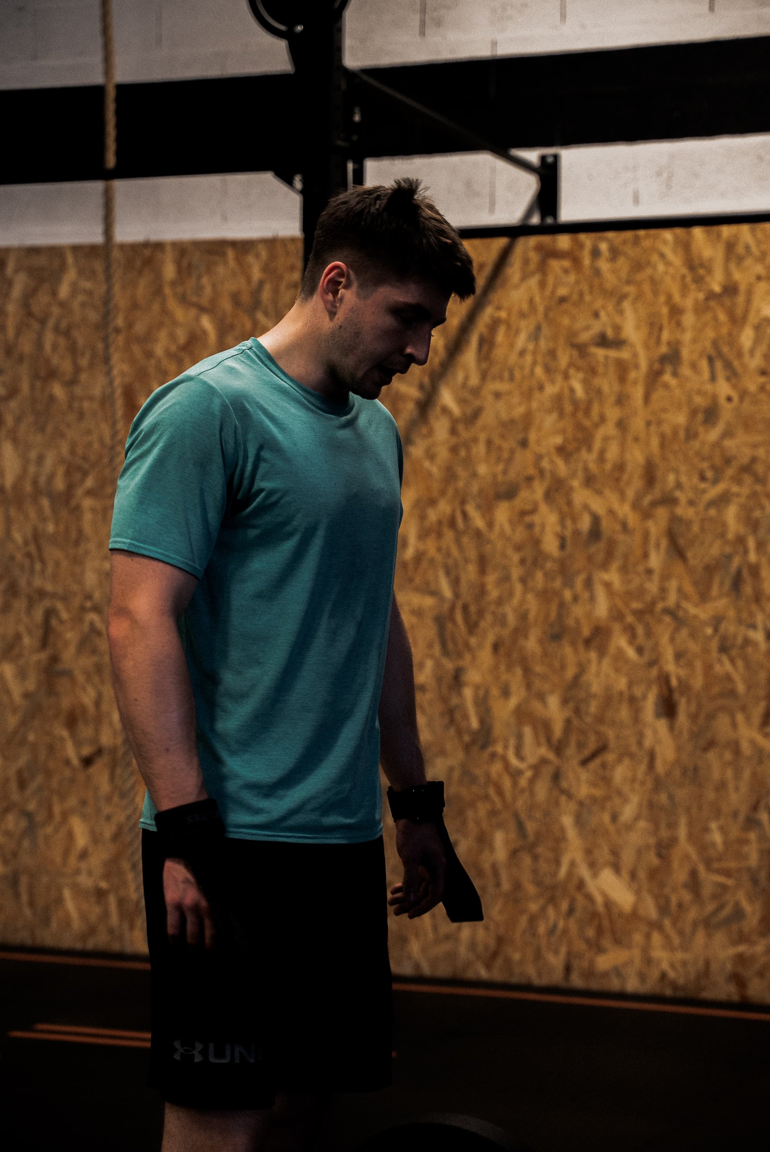 Un homme en t-shirt bleu et shorts noirs dans une salle de sport, se préparant à faire de l'exercice, avec un mur en bois en arrière-plan.